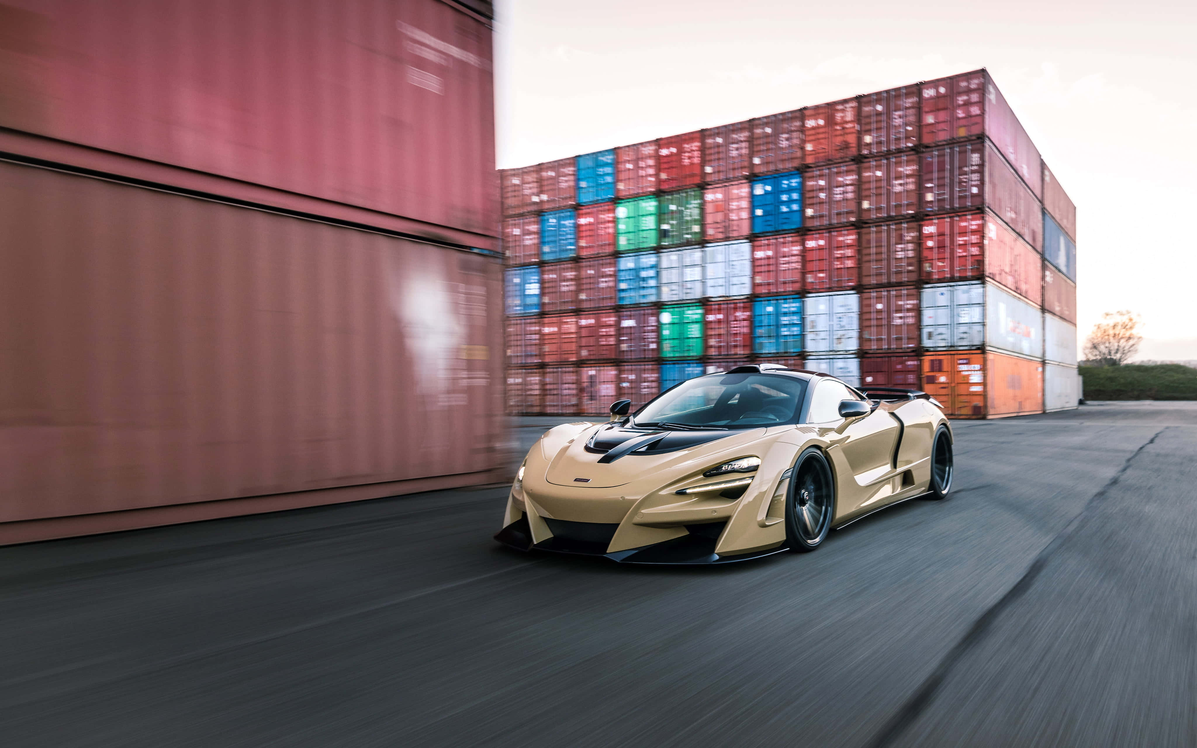 Mclaren 720s 桌布 壁紙 — 金色麥拉倫超級跑車在動態模糊鏡頭中高速駛過堆疊的貨運貨櫃。