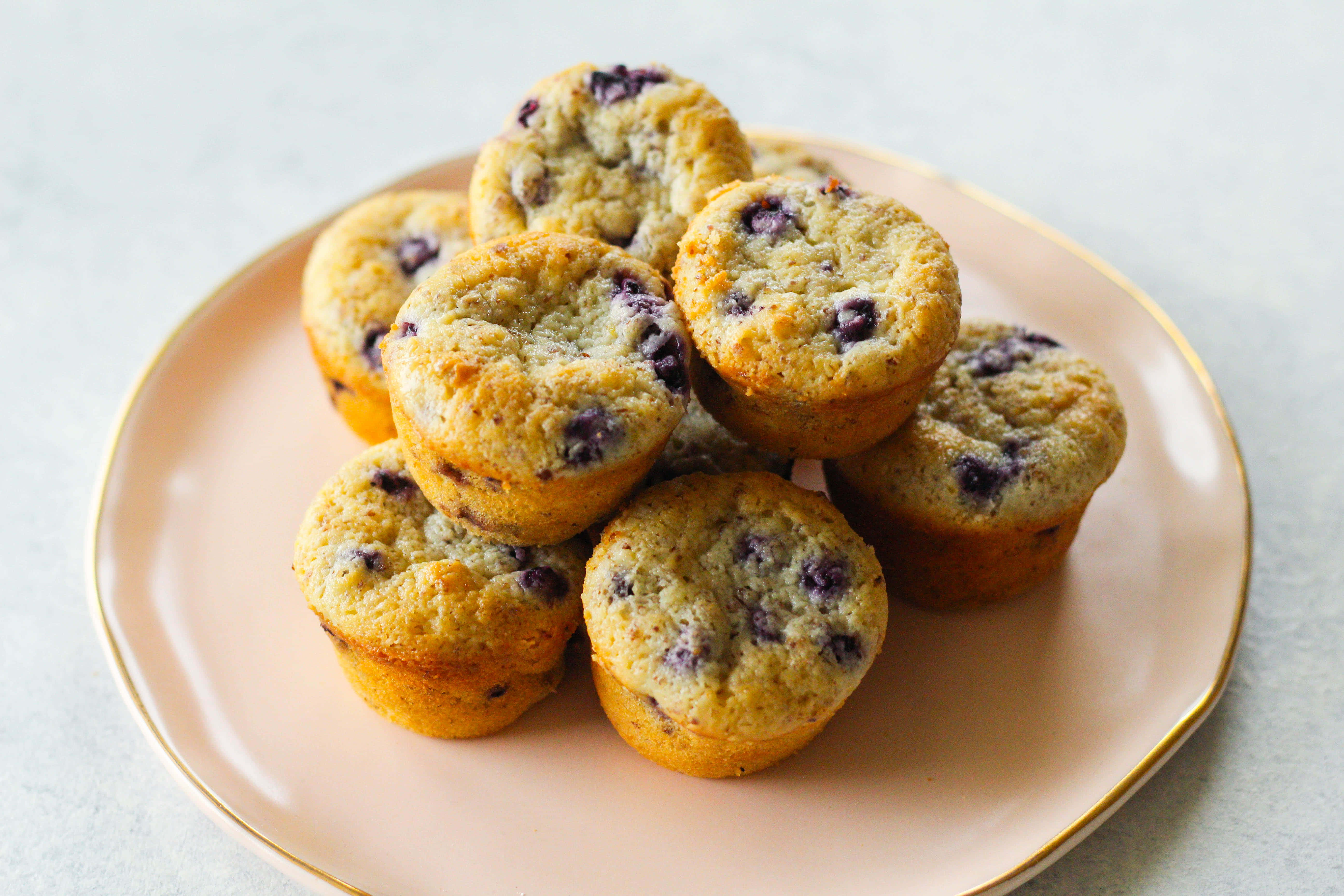 Blueberry Muffins 桌布 壁紙 — 金藍莓馬芬高高堆放在柔軟的粉紅色金邊盤子上，背景為淺灰色。