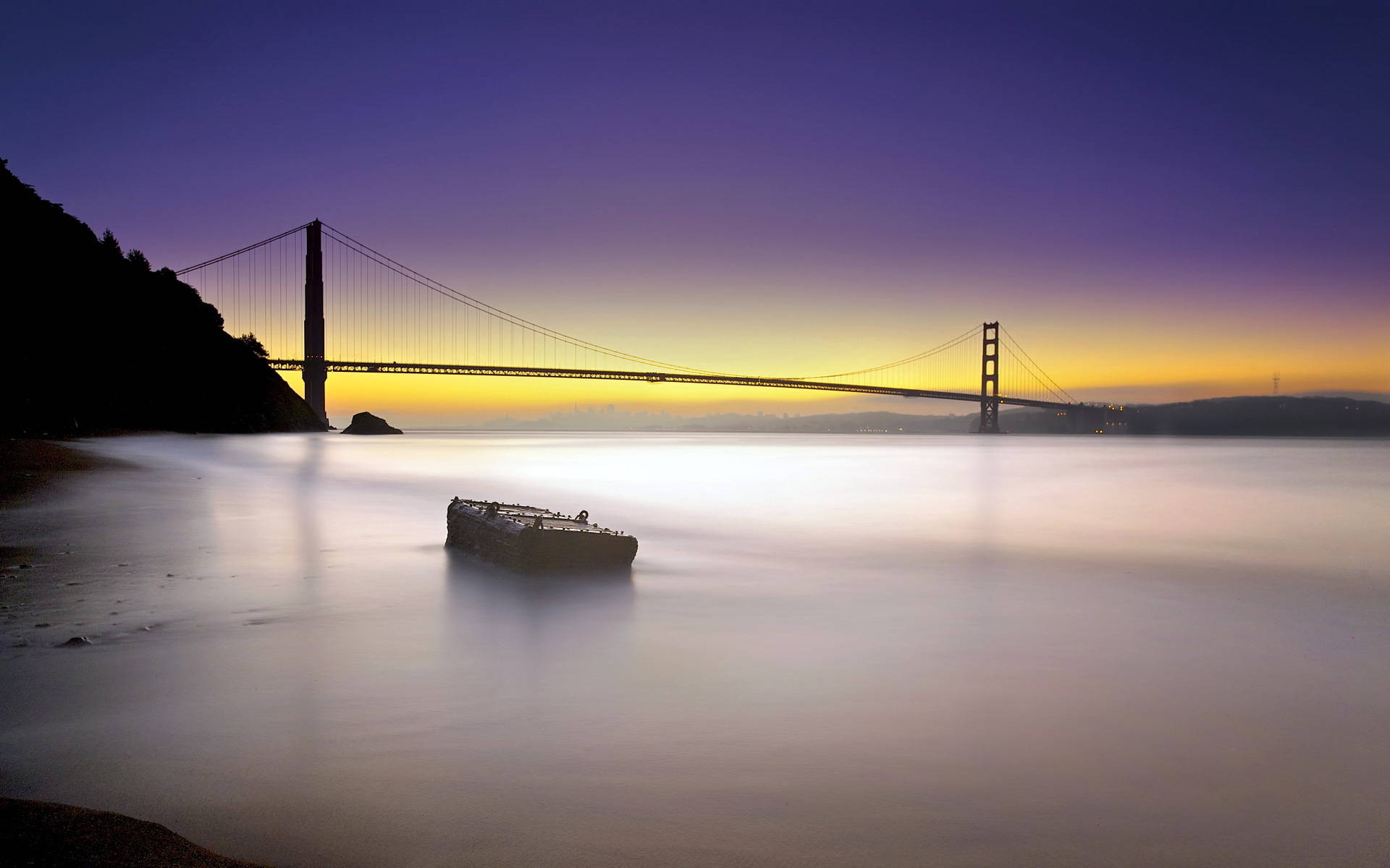 San Francisco HD 桌布 壁紙 — 金門大橋在充滿活力的紫色和橙色日落中形成剪影，一艘幽靈船漂浮在平靜的水面上。