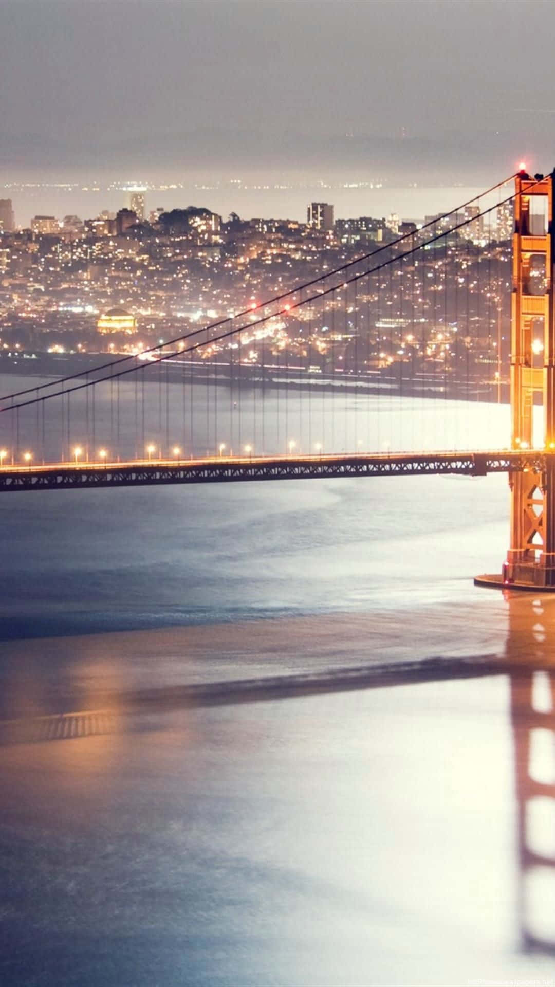 桌布 壁紙 — 金門大橋在夜晚溫暖地發光，平靜的水面上遠處的城市燈光閃爍。