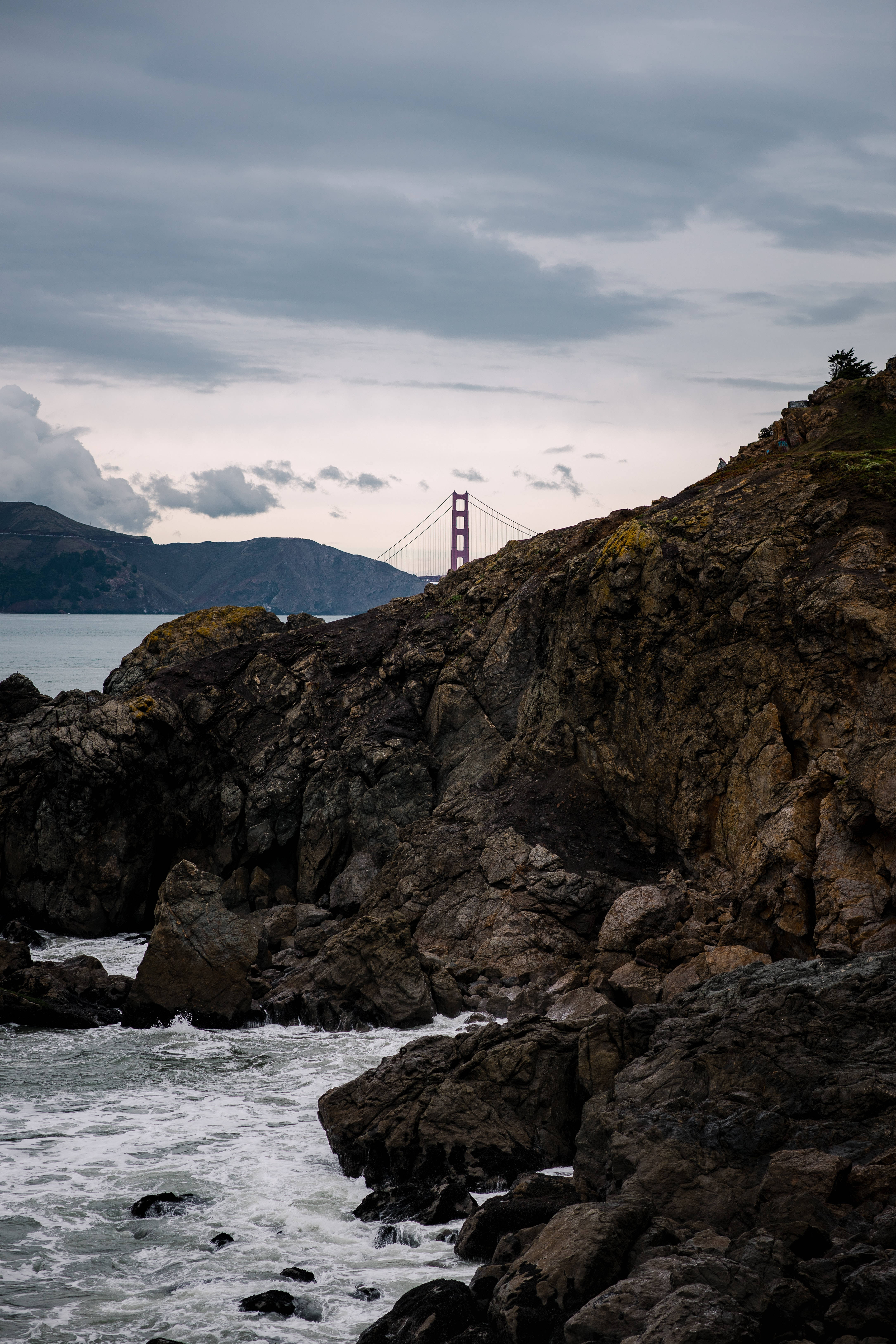 Rock 桌布 壁紙 — 金門大橋在崎嶇懸崖後方遠處矗立，海浪拍打著暗色岩石，天空陰沉灰暗。