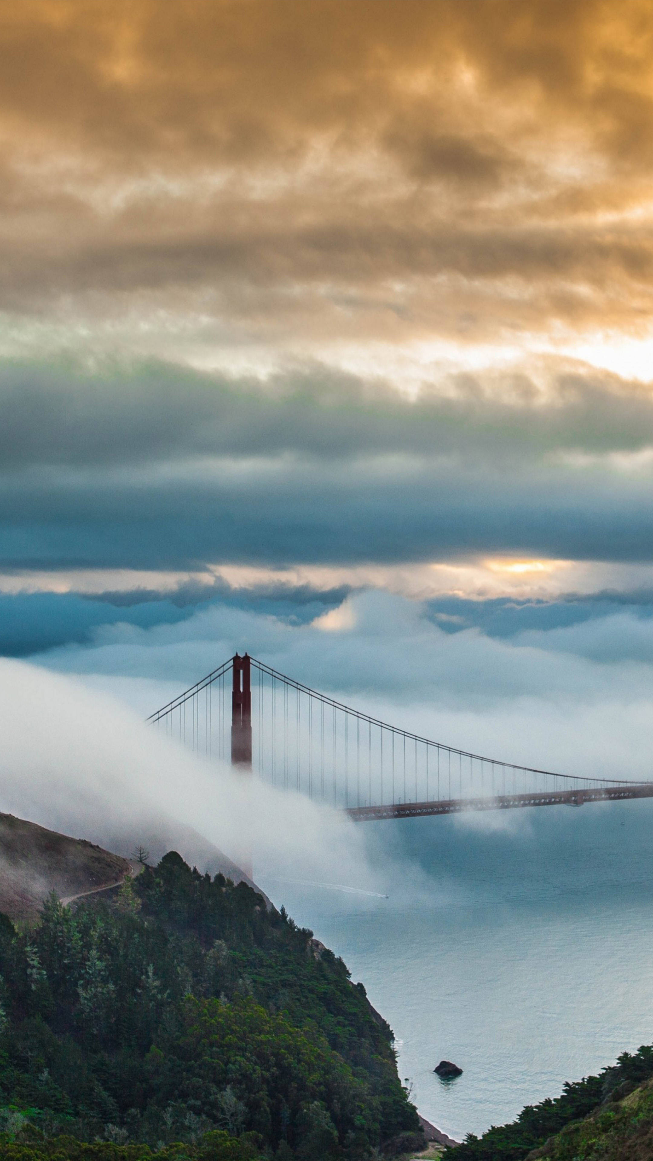 Clouds Aesthetics 桌布 壁紙 — 金門大橋在戲劇性的日出天空下，從薄霧中浮現，伴隨著層層雲朵和綠色山坡。