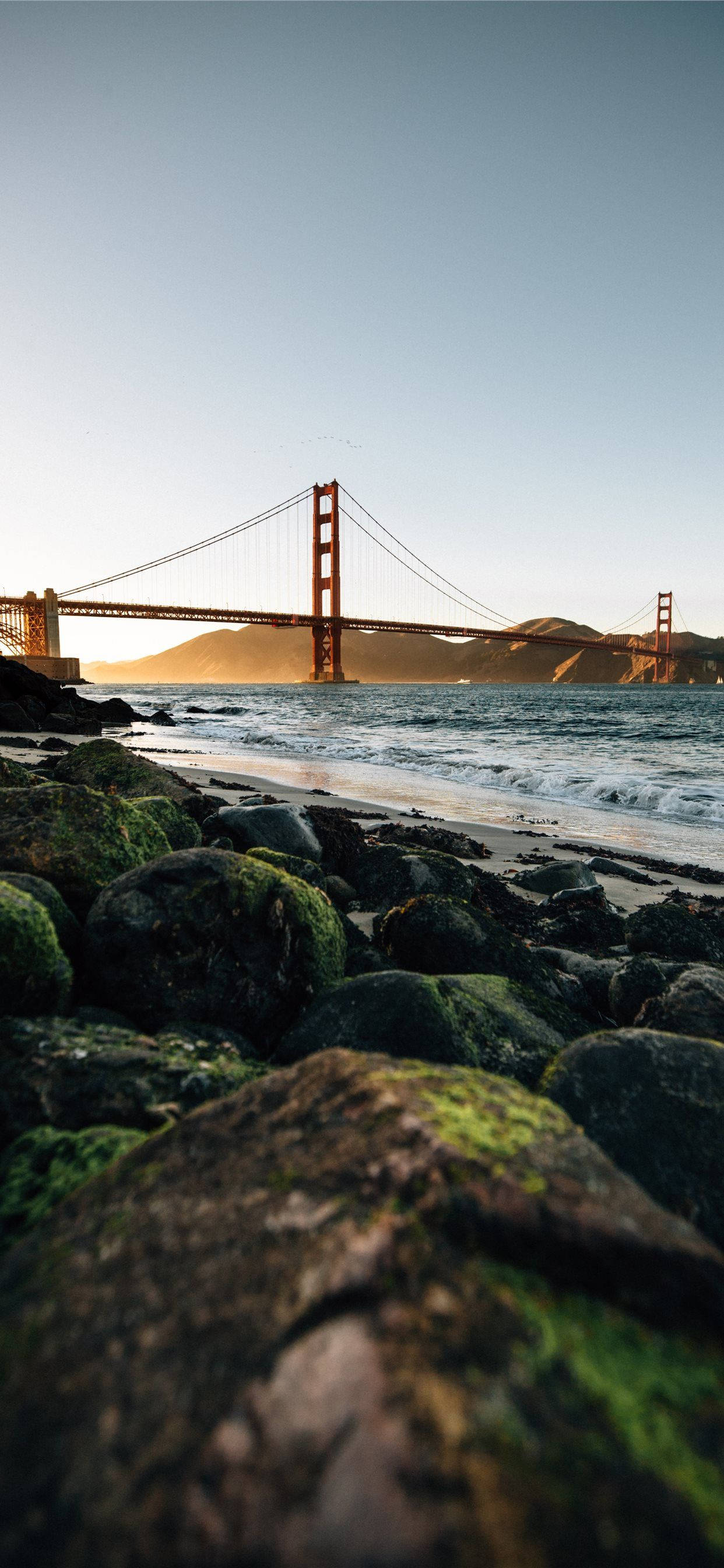 san francisco iPhone 桌布 壁紙 — 金門大橋在日落時橫跨海灣，前景有長滿苔蘚的岩石，下方是平靜的水面。