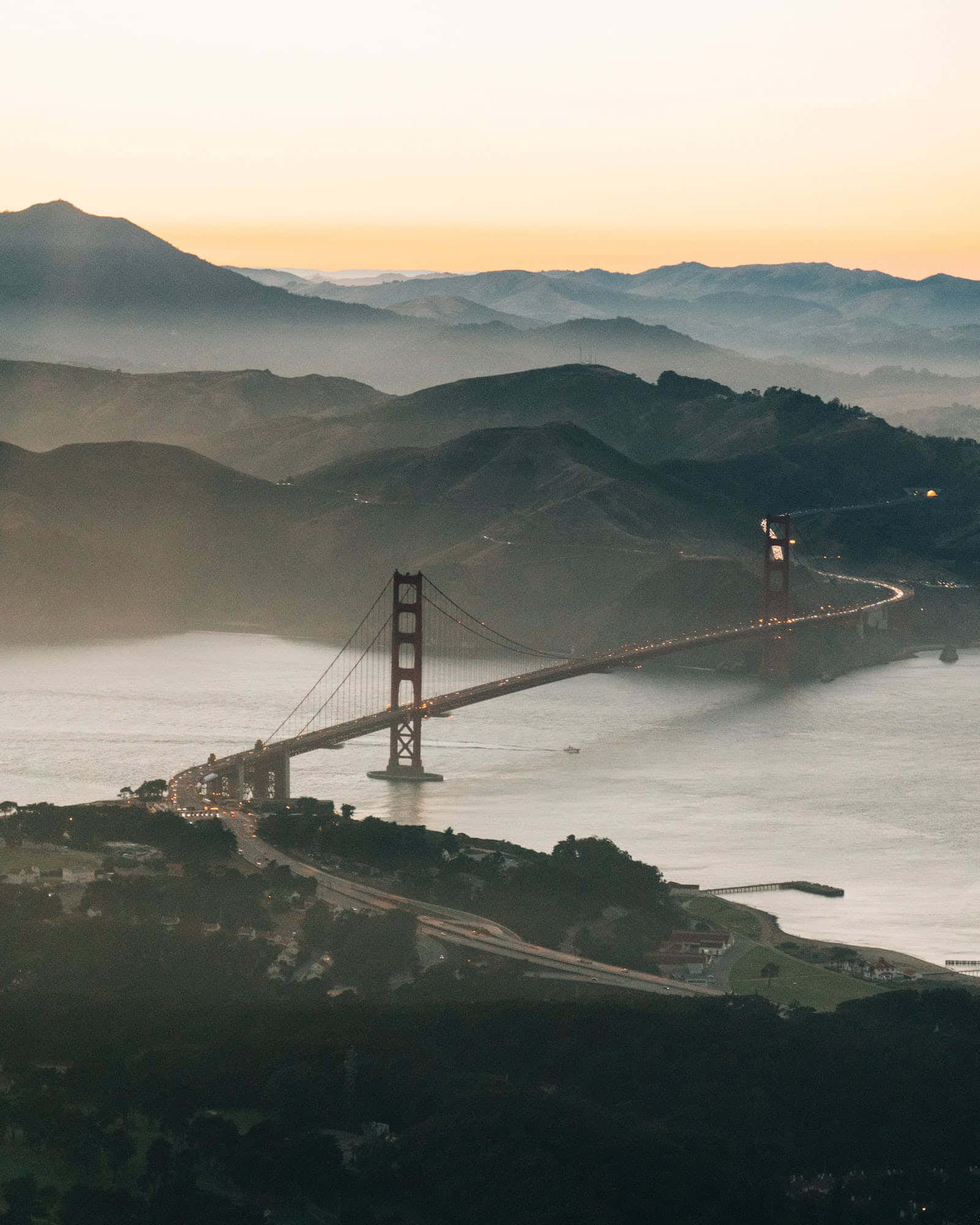San Francisco Fog 桌布 壁紙 — 金門大橋在日落時閃耀，下方有霧氣籠罩的山丘和寧靜的水面。