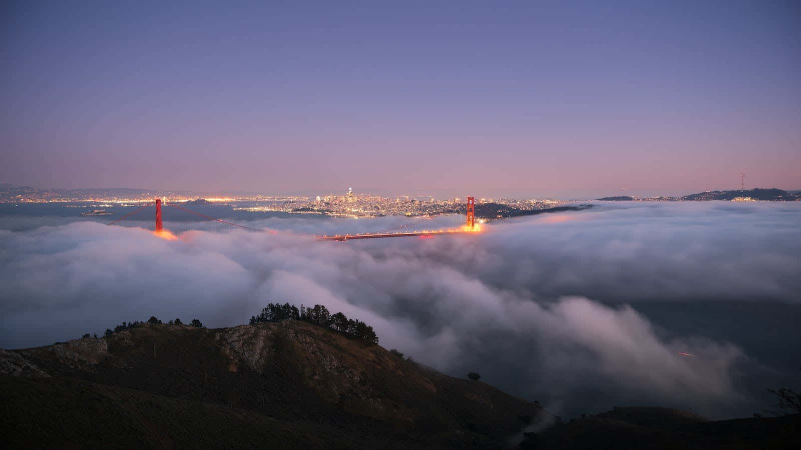 San Francisco Fog 桌布 壁紙 — 金門大橋在暮色中從雲海中浮現，下方城市燈光閃爍。