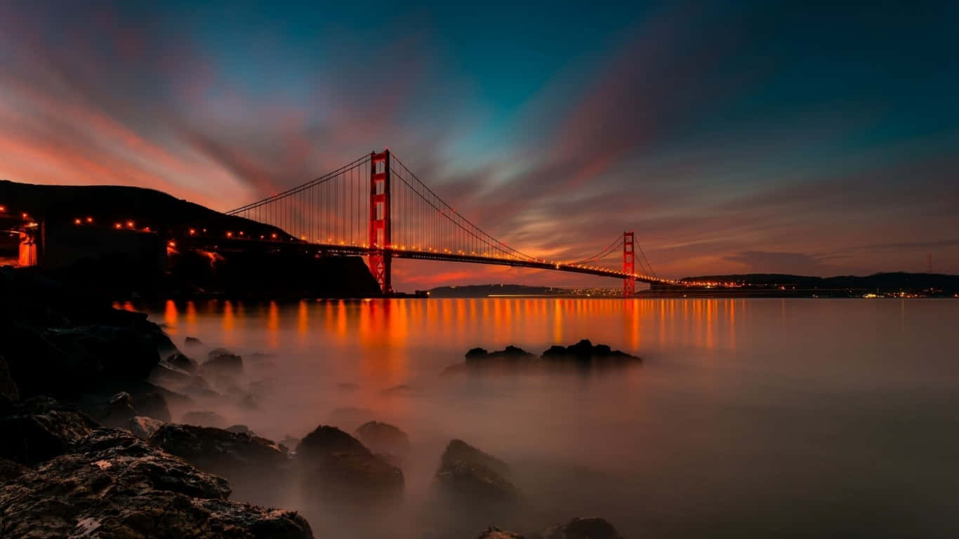 1366x768 San Francisco 桌布 壁紙 — 金門大橋在暮色天空、條紋雲朵和倒映燈光的平靜水面襯托下，散發出火熱的橙色光芒。