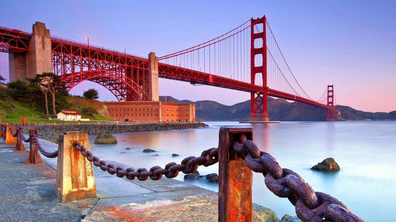 1366x768 San Francisco 桌布 壁紙 — 金門大橋在暮色天空下泛著紅光，生鏽的鐵鏈框住了前景平靜的海灣水面。