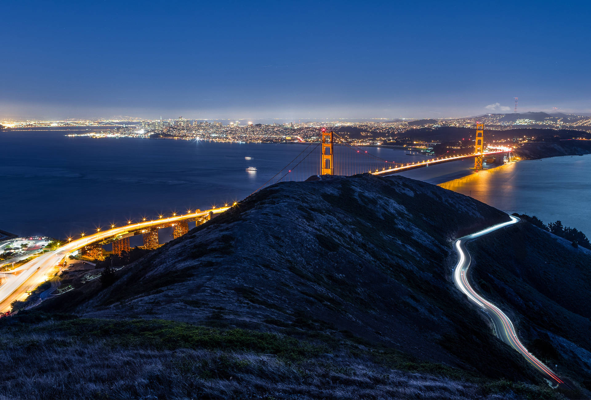 lights 桌布 壁紙 — 金門大橋在暮色天空下溫暖地發光，光跡蜿蜒沿著俯瞰舊金山灣的黑暗山坡而下。