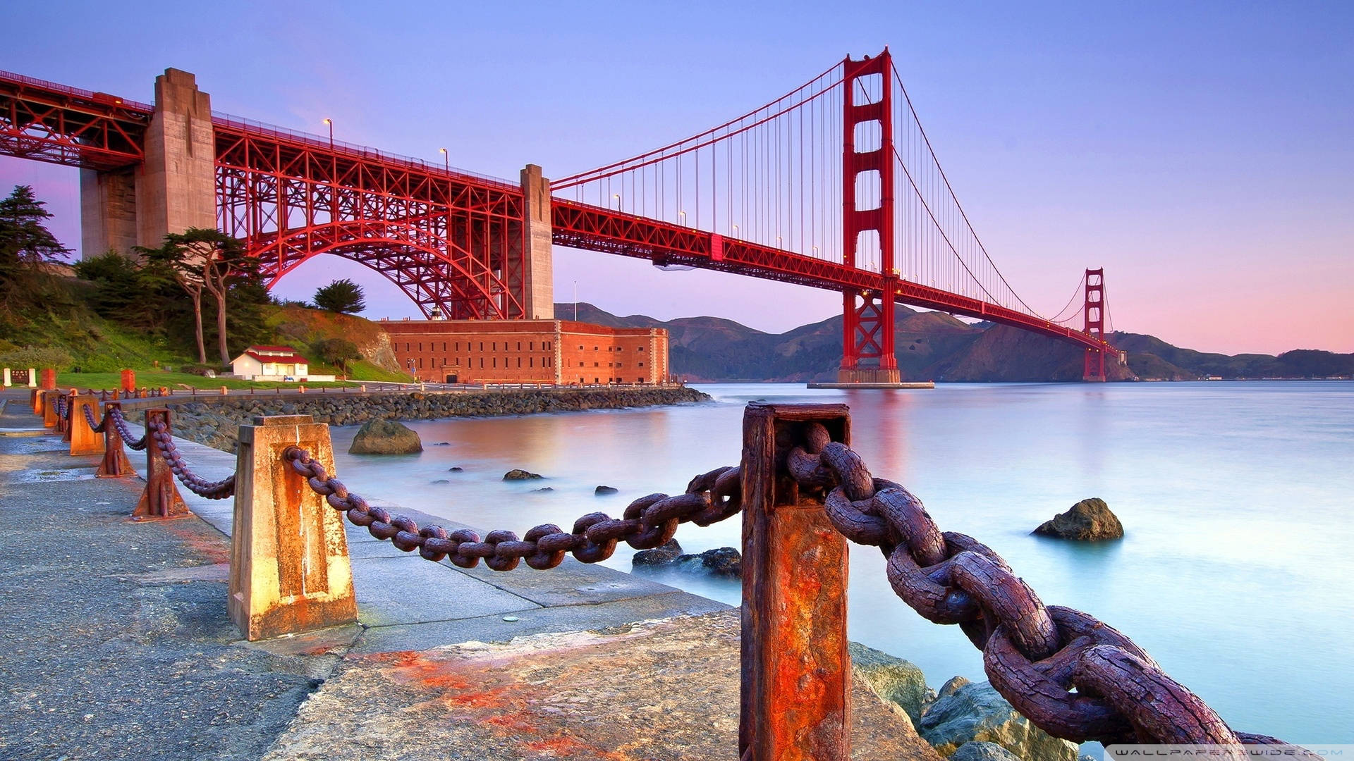 golden gate bridge 桌布 壁紙 — 金門大橋在柔和的紫色日落映襯下發出紅光，鏽蝕的鐵鏈沿著石碼頭排列，俯瞰平靜的水面。