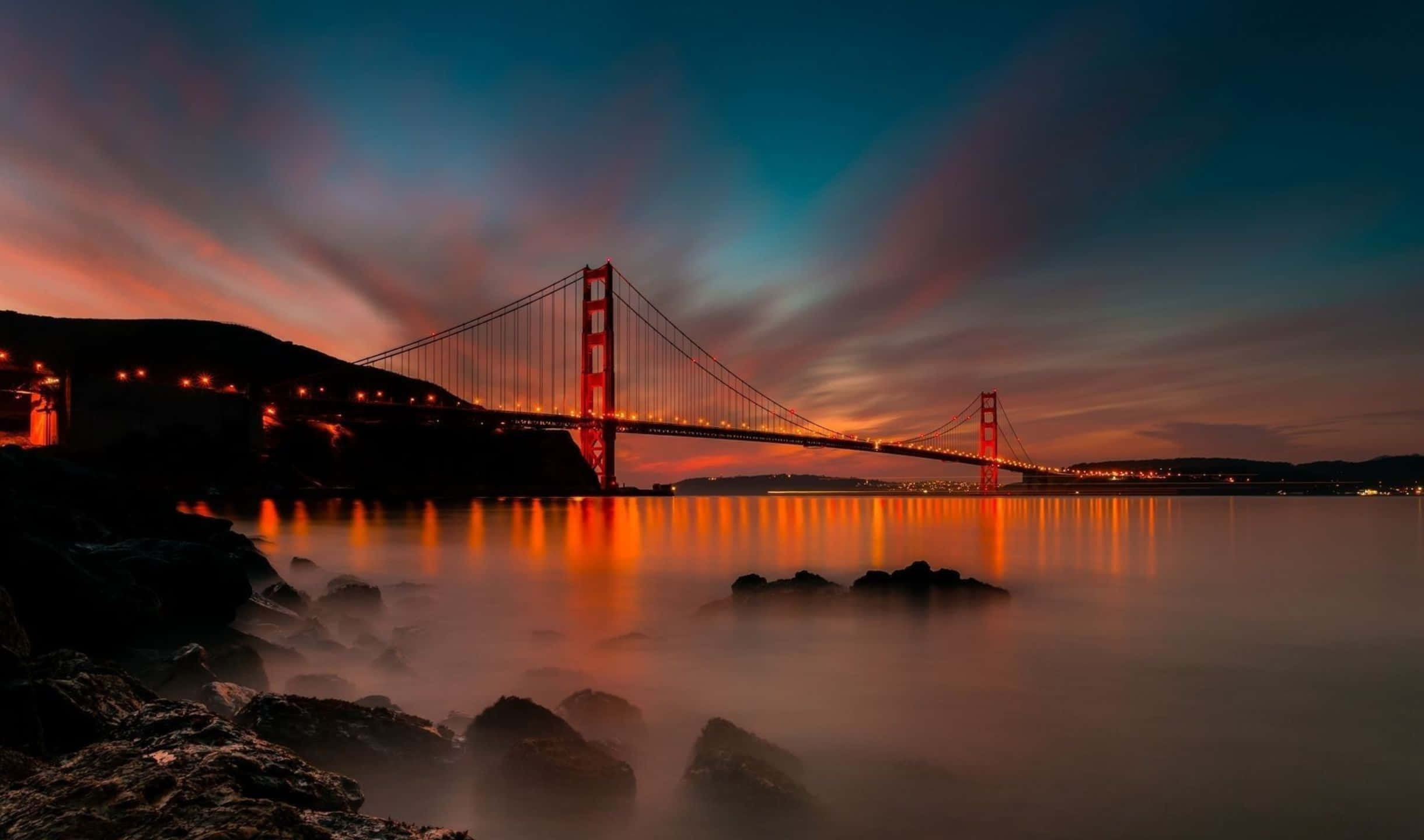 2440x1440 San Francisco 桌布 壁紙 — 金門大橋在橙藍交織的暮色天空中溫暖地發光，其燈光映照在霧氣瀰漫的水面上。