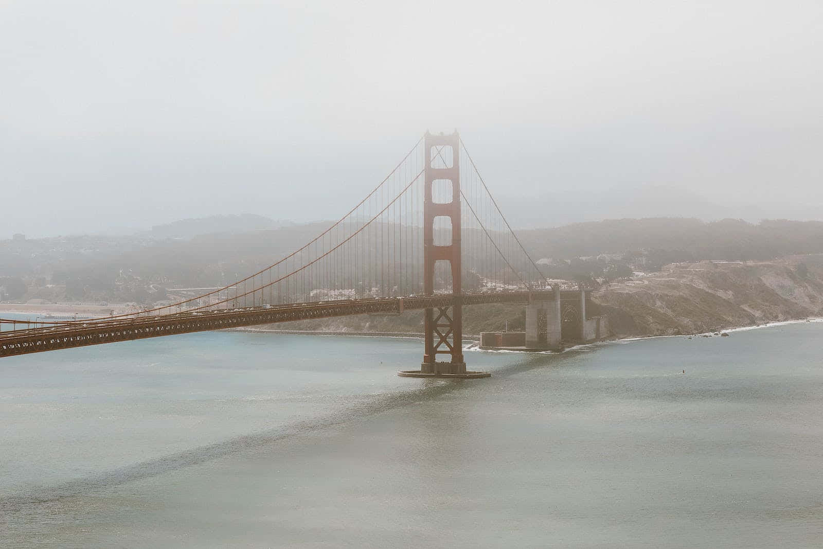 San Francisco Fog 桌布 壁紙 — 金門大橋在濃霧中浮現於平靜的水面上，背景是朦朧的山丘。
