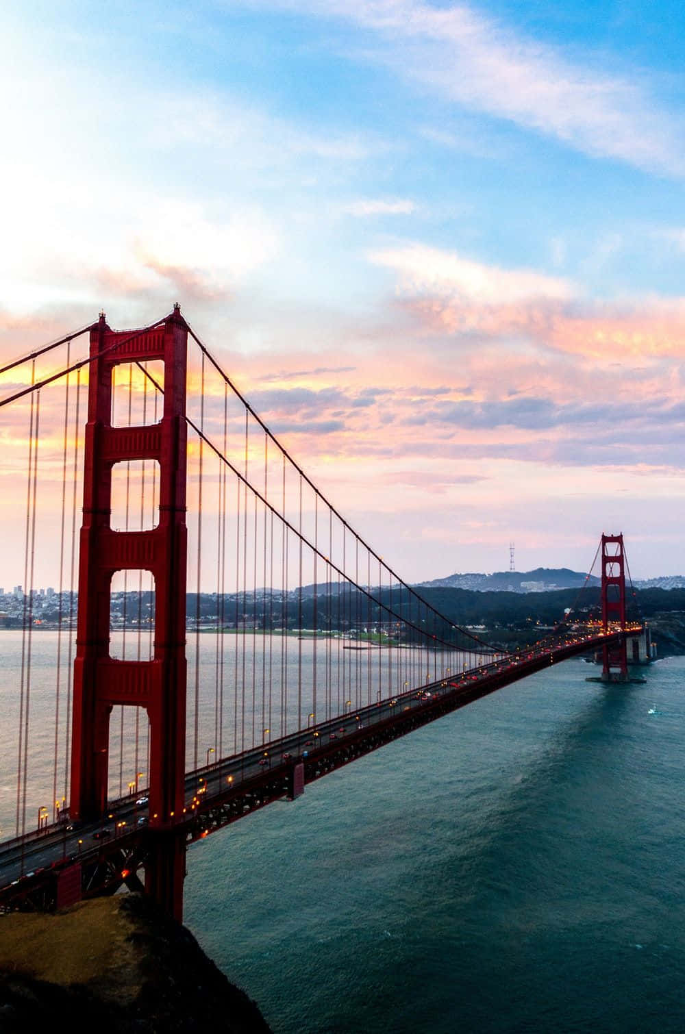 bridge 桌布 壁紙 — 金門大橋在粉彩色的日落天空下泛著紅光，汽車在平靜的水面上沿著橋樑行駛。