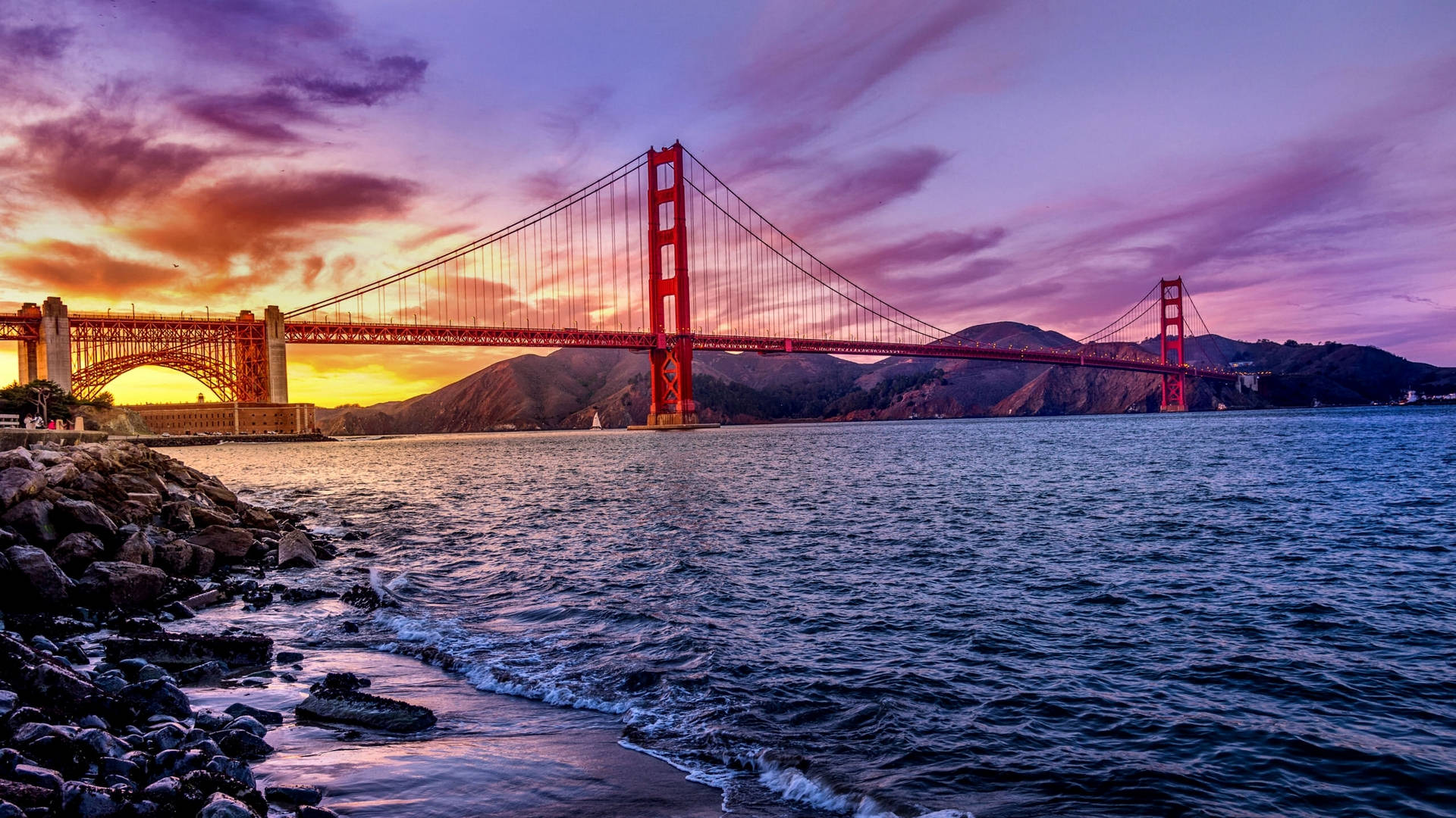 San Francisco HD 桌布 壁紙 — 金門大橋在紫色和橙色鮮豔的日落天空下，在翻騰的海浪和岩石海岸上閃耀著紅光。