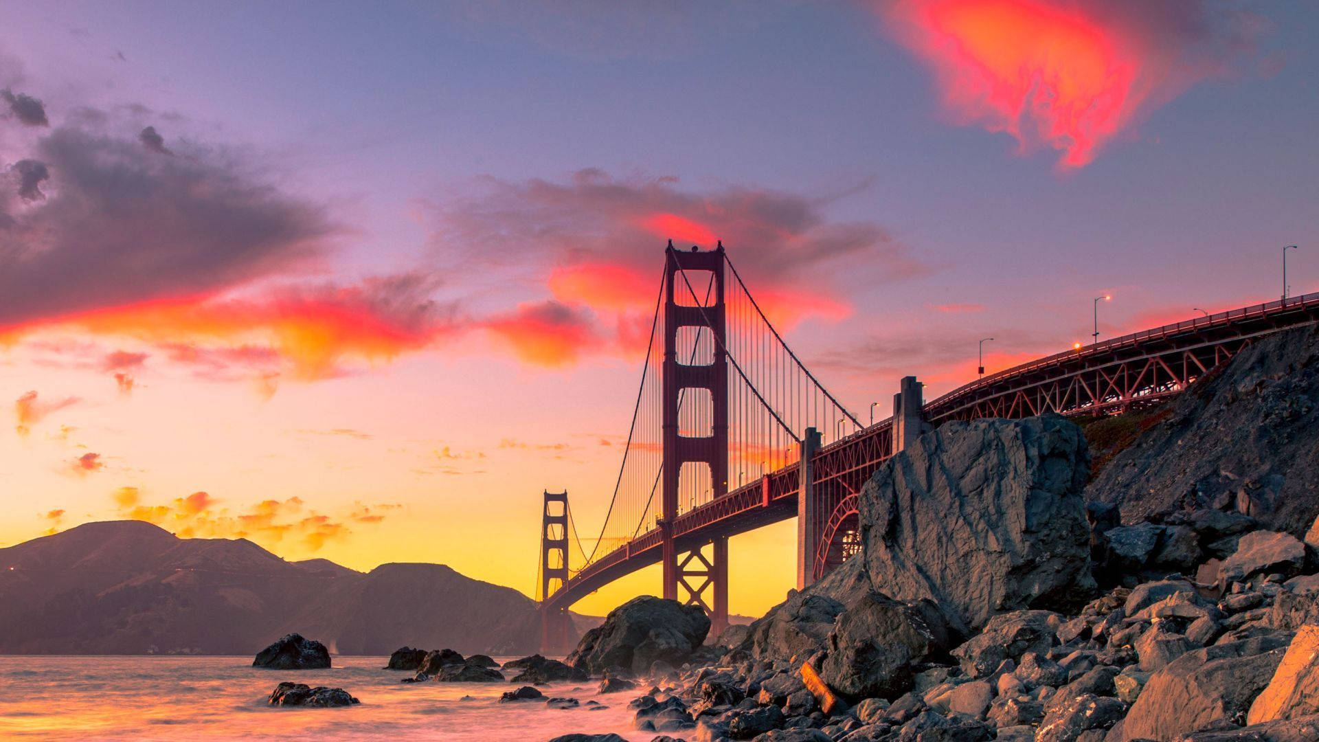 San Francisco HD 桌布 壁紙 — 金門大橋在紫色天空、岩石前景和寧靜水面的襯托下，閃耀著火熱的日落色調。