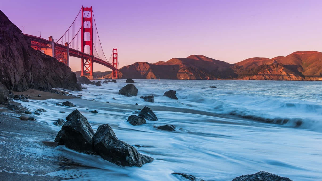 1366x768 San Francisco 桌布 壁紙 — 金門大橋在紫色日落映襯下發出紅光，海浪平穩地流過霧氣籠罩的海岸線上的岩石。