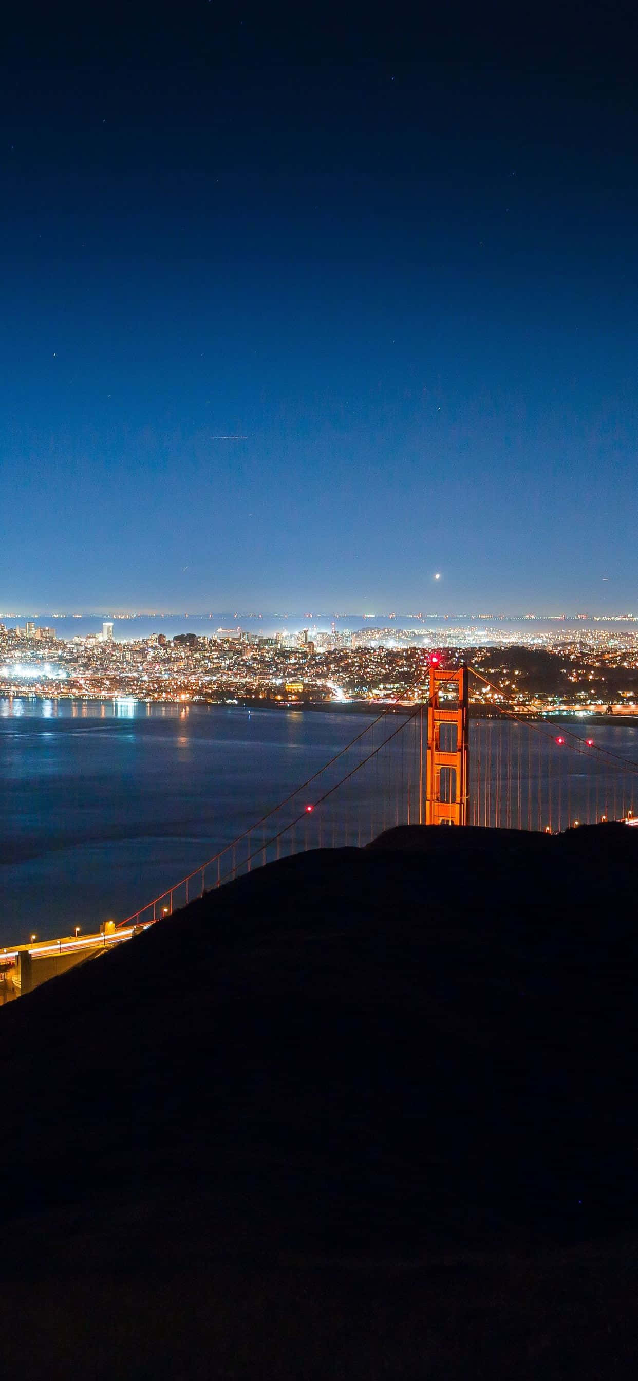 bridge 桌布 壁紙 — 金門大橋在繁星點點的夜空下溫暖地發光，俯瞰著舊金山閃爍的城市景觀和黑暗的海灣水域。