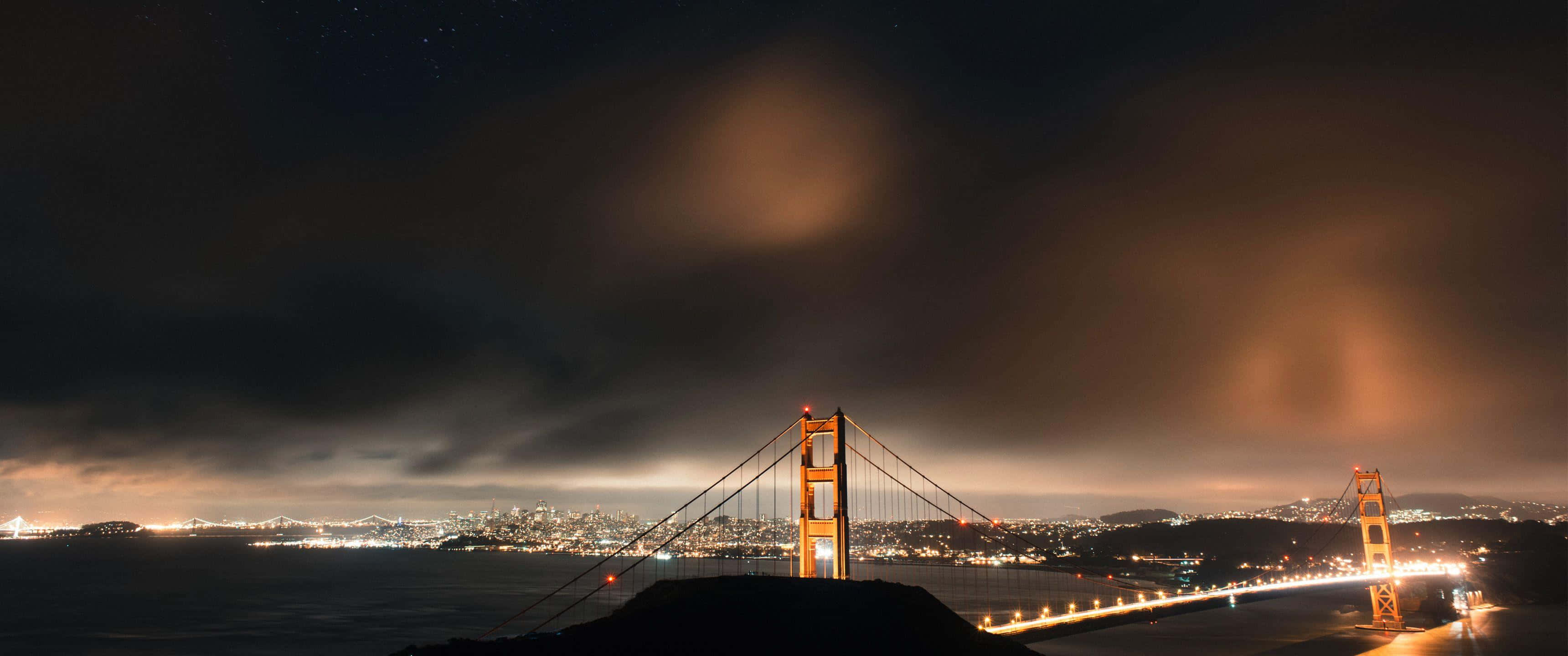 Ultra Wide 桌布 壁紙 — 金門大橋在繁星點點的夜空下溫暖地發光，下方有戲劇性的雲彩和城市燈光。