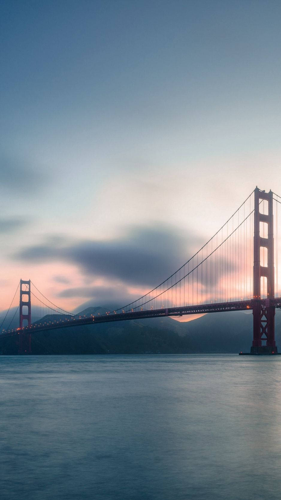 san francisco iPhone 桌布 壁紙 — 金門大橋在黃昏時分發出光芒，上方是柔和的粉紅色和藍色天空，下方是平靜的水面。