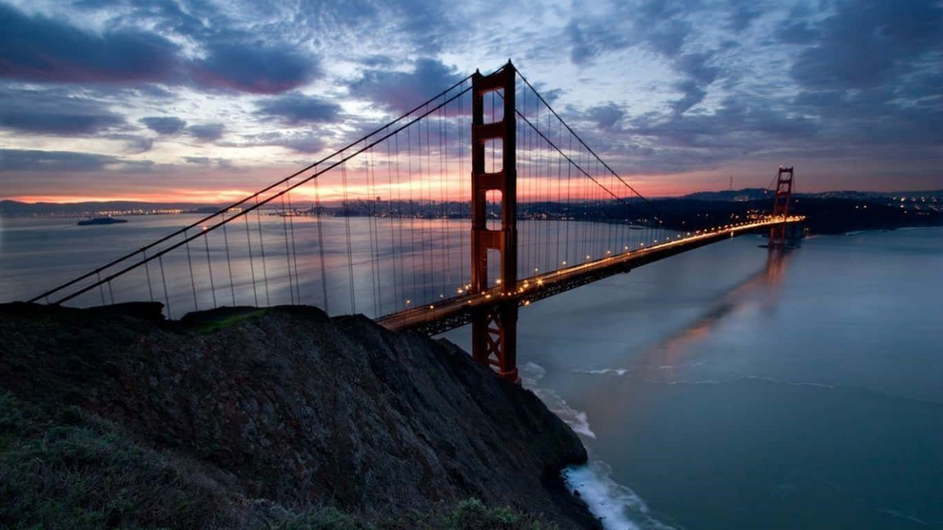 1366x768 San Francisco 桌布 壁紙 — 金門大橋在黃昏時分發出溫暖的光芒，映襯著紫色和橙色的天空，下方是平靜的水面。