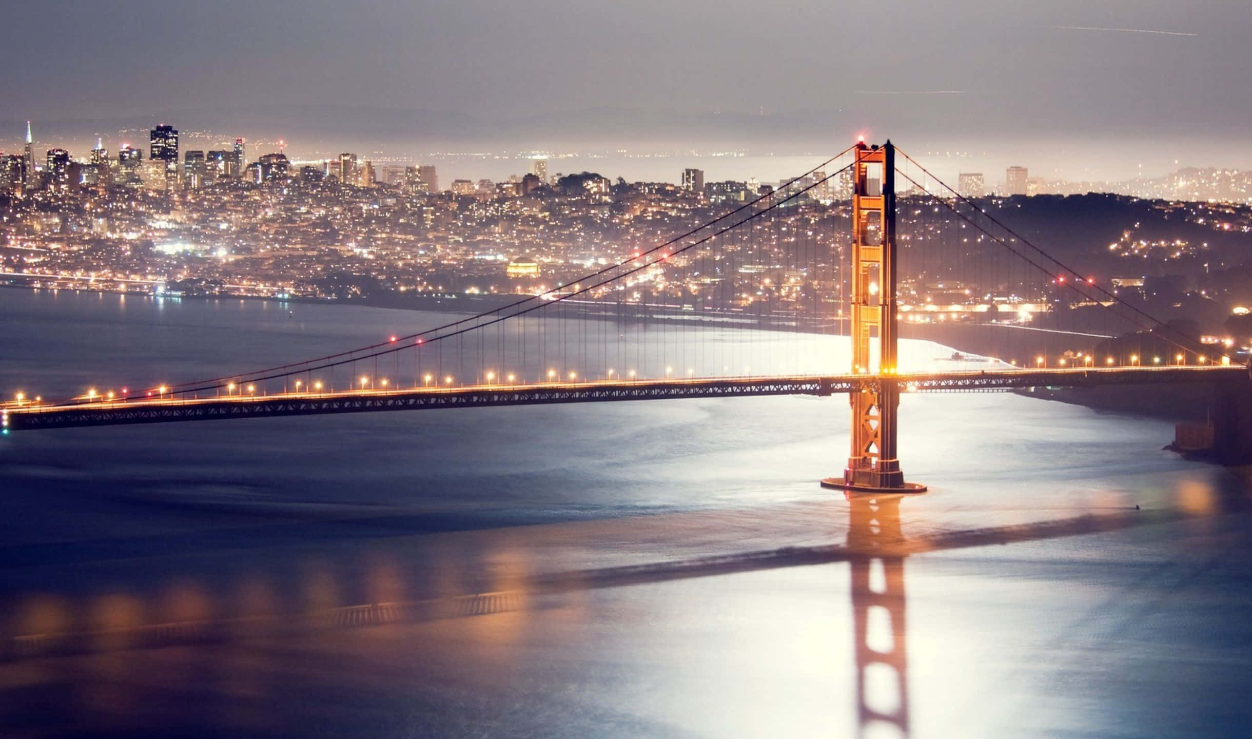 2440x1440 San Francisco 桌布 壁紙 — 金門大橋在黃昏時溫暖地發光，城市燈光在其標誌性塔樓下的平靜水面上反射。