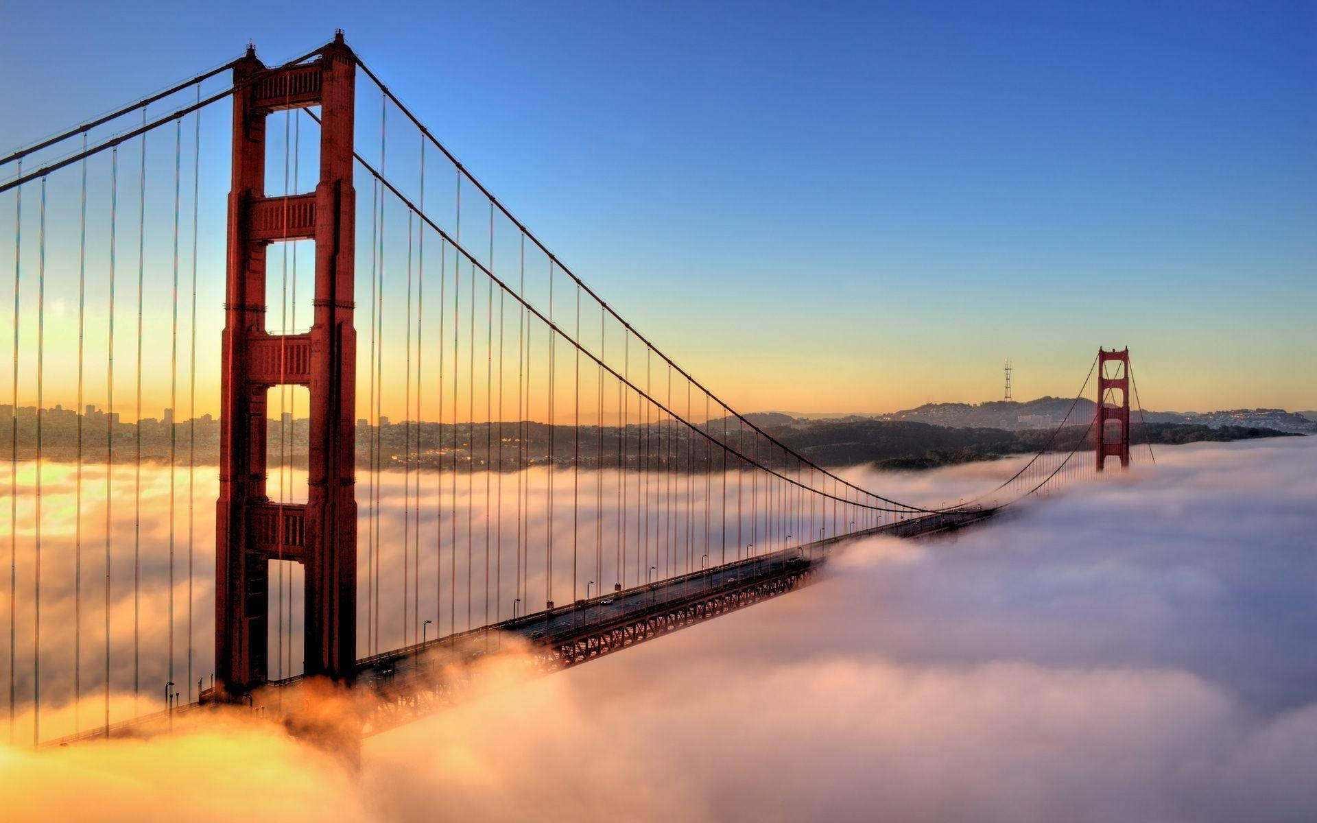 San Francisco HD 桌布 壁紙 — 金門大橋從雲海中浮現，日出時溫暖的橙色光芒灑落在它的橋塔上，映襯著清澈的藍天。