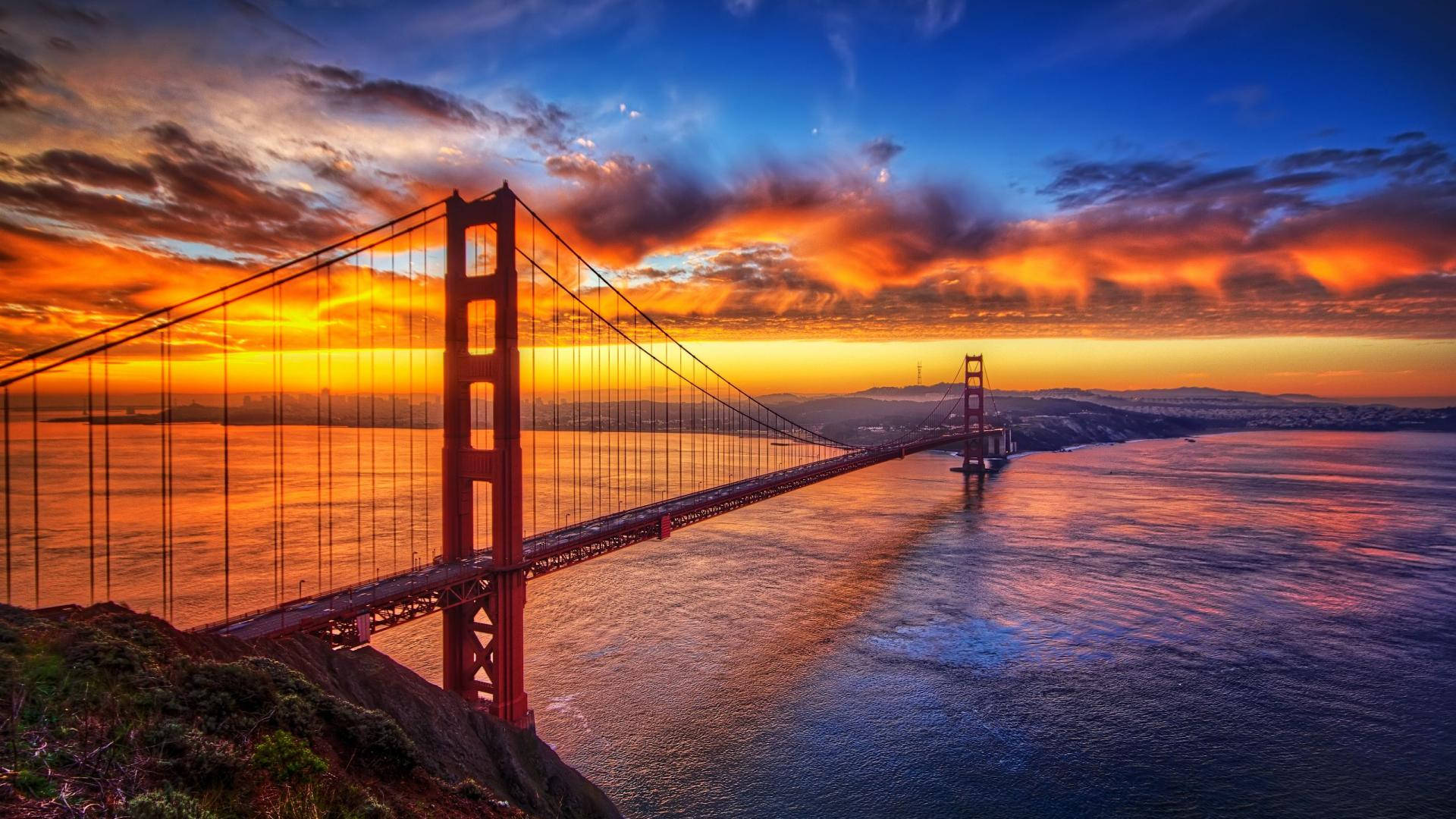 San Francisco 桌布 壁紙 — 金門大橋沐浴在熾熱的日落色調中，橫跨舊金山灣，伴隨著戲劇性的雲彩和閃爍的水面。
