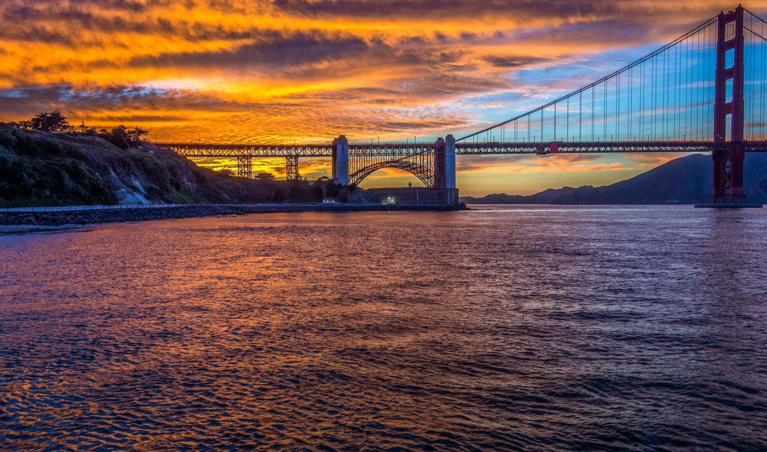 2440x1440 San Francisco 桌布 壁紙 — 金門大橋沐浴在熾熱的日落色調中，下方是漣漪的水面，天空有戲劇性的橙色和紫色雲彩。