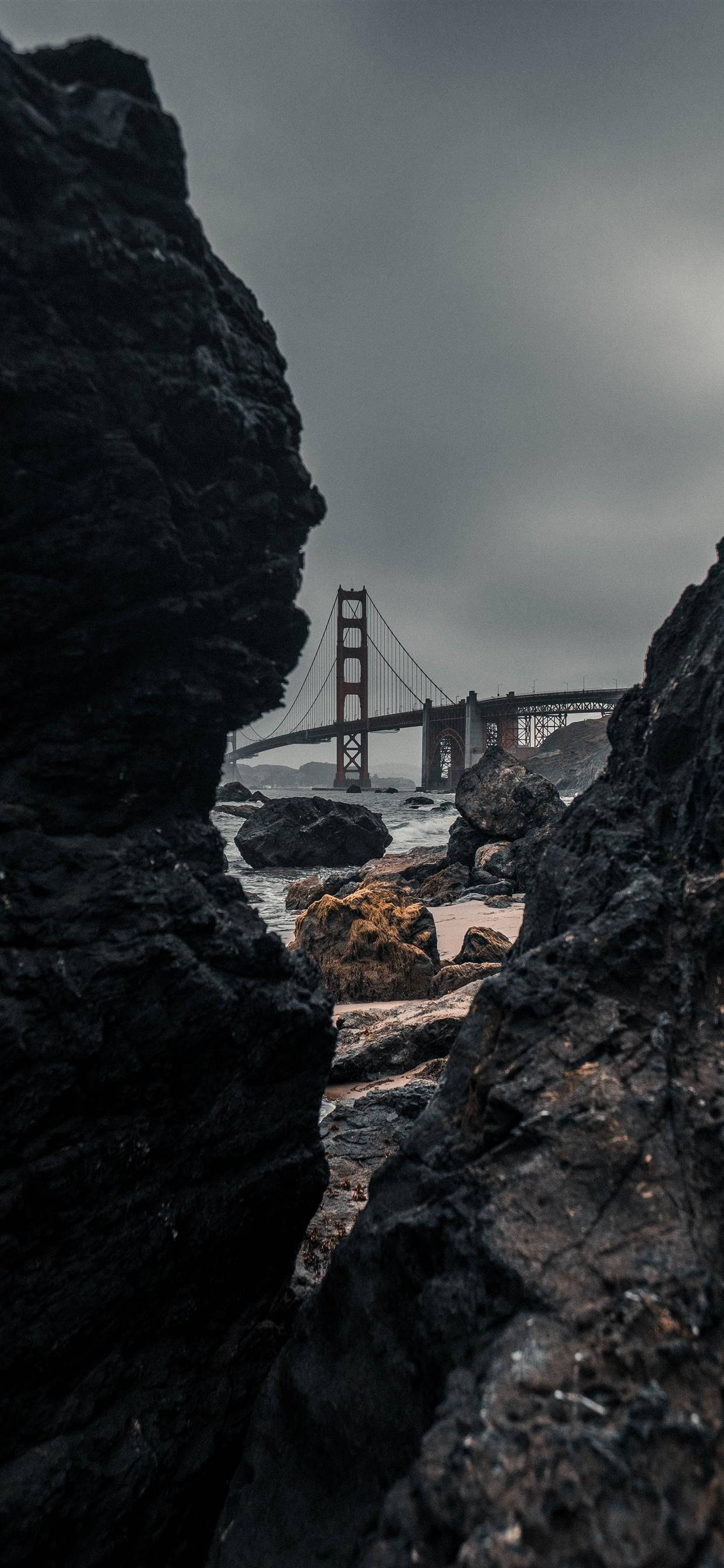 san francisco iPhone 桌布 壁紙 — 金門大橋透過暗黑崎嶇的岩石觀看，陰沉灰暗的天空下波濤洶湧。