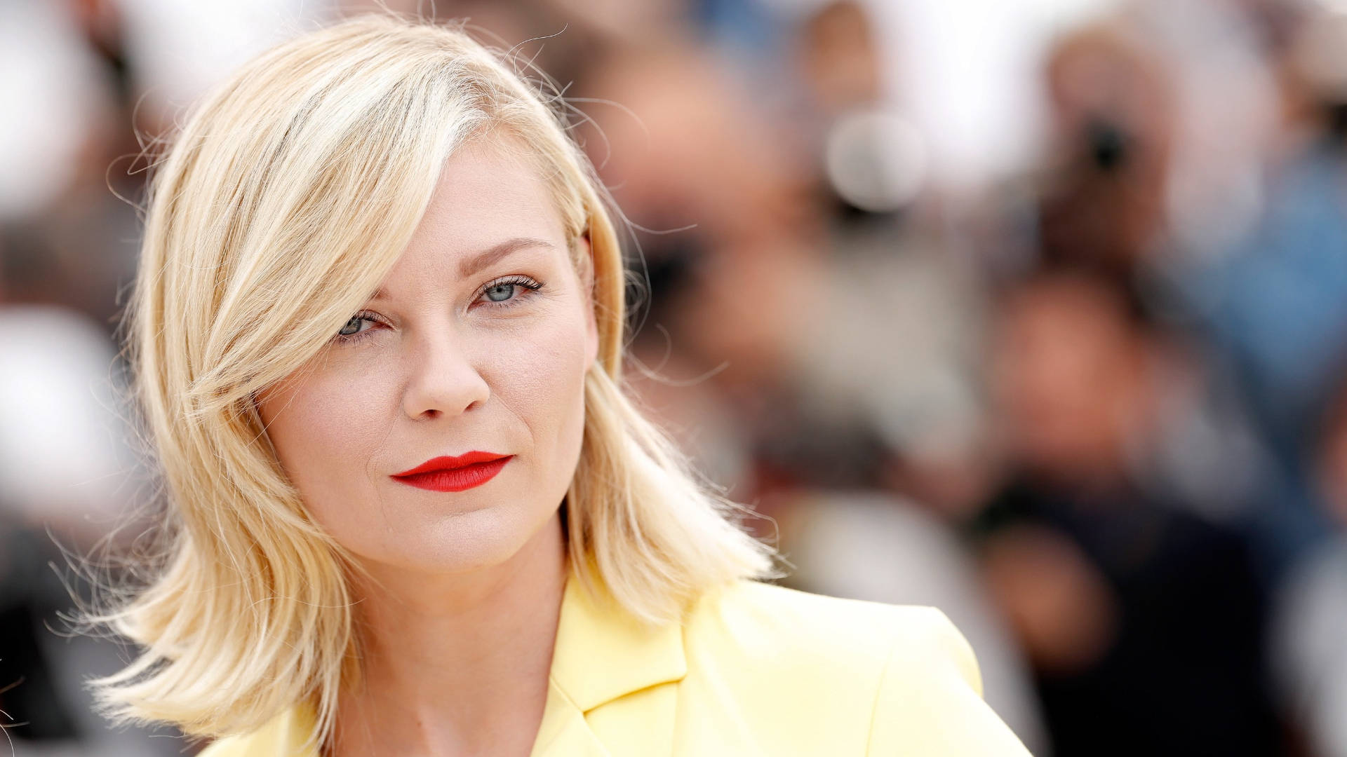 Kirsten Dunst 桌布 壁紙 — 金髮女子塗紅色唇膏身穿黃色西裝外套，背景為模糊人群的特寫肖像。