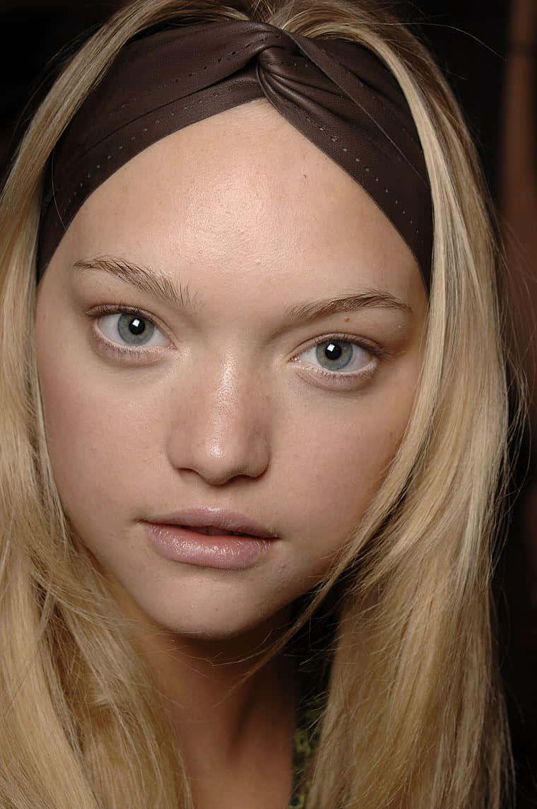 Gemma Ward 桌布 壁紙 — 金髮模特兒的近距離肖像，擁有引人注目的藍色眼睛，戴著棕色打結頭帶，背景為深色。