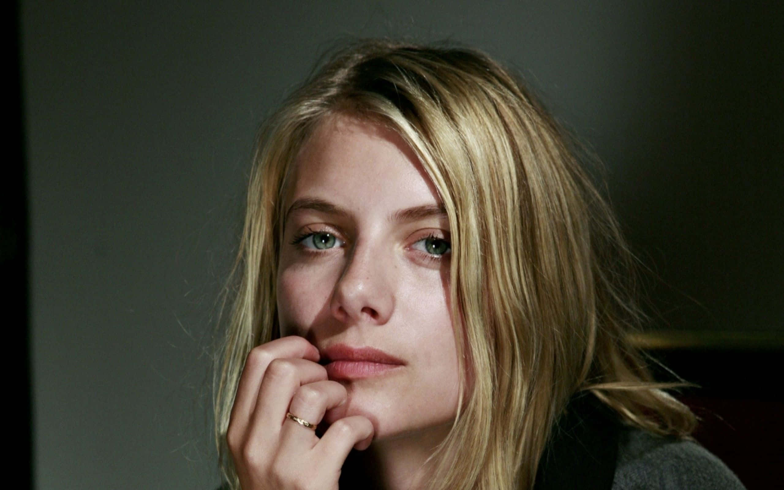Mélanie Laurent 桌布 壁紙 — 金髮綠眼女子的特寫肖像，她將下巴擱在手上，背景是柔和的灰色。