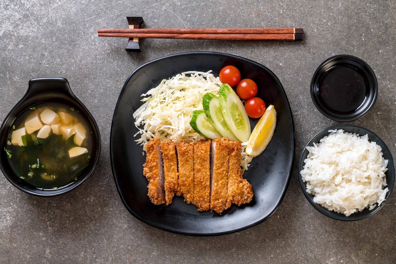 Food Photography 桌布 壁紙 — 金黃色切塊炸豬排放在黑色盤子上，配上高麗菜絲、小番茄、小黃瓜、白飯和味噌湯。