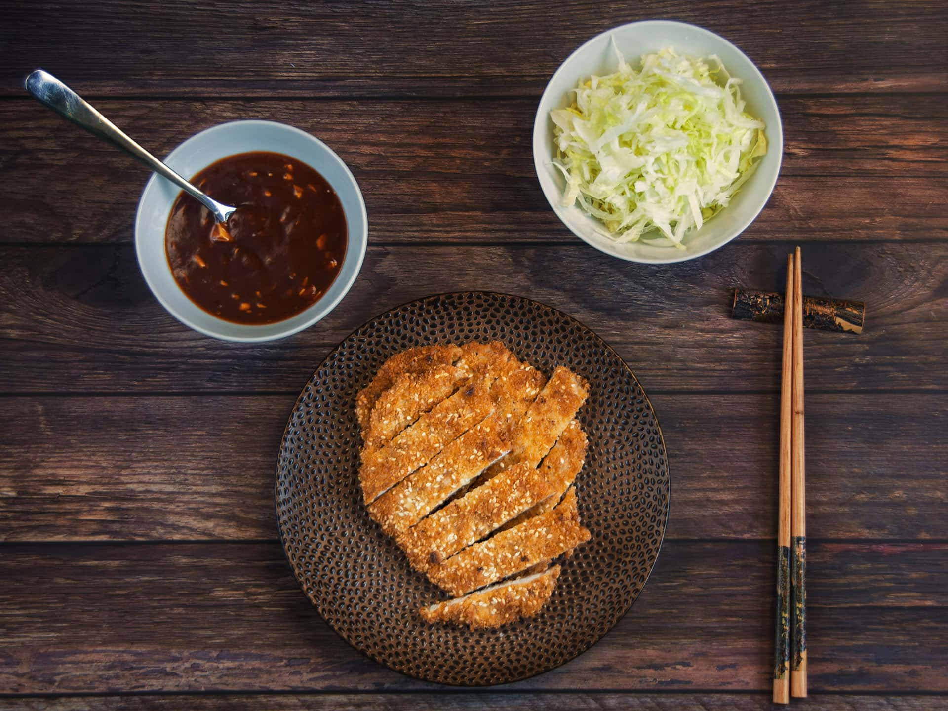Food Photography 桌布 壁紙 — 金黃色切片炸豬排放在有紋理的盤子上，搭配高麗菜和醬汁，置於深色木桌上。