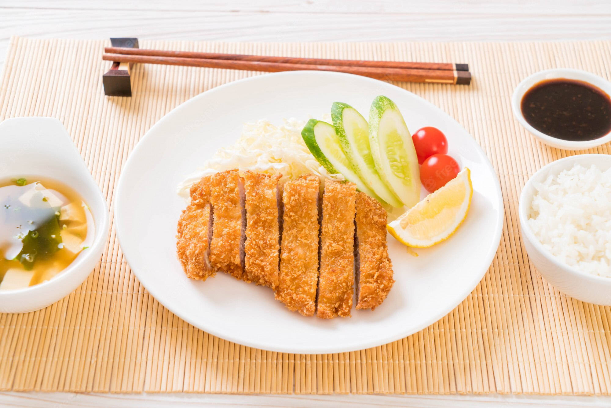 Food Photography 桌布 壁紙 — 金黃色切片炸豬排放在白色盤子上，搭配米飯、小黃瓜、番茄和醬汁，置於竹蓆上。