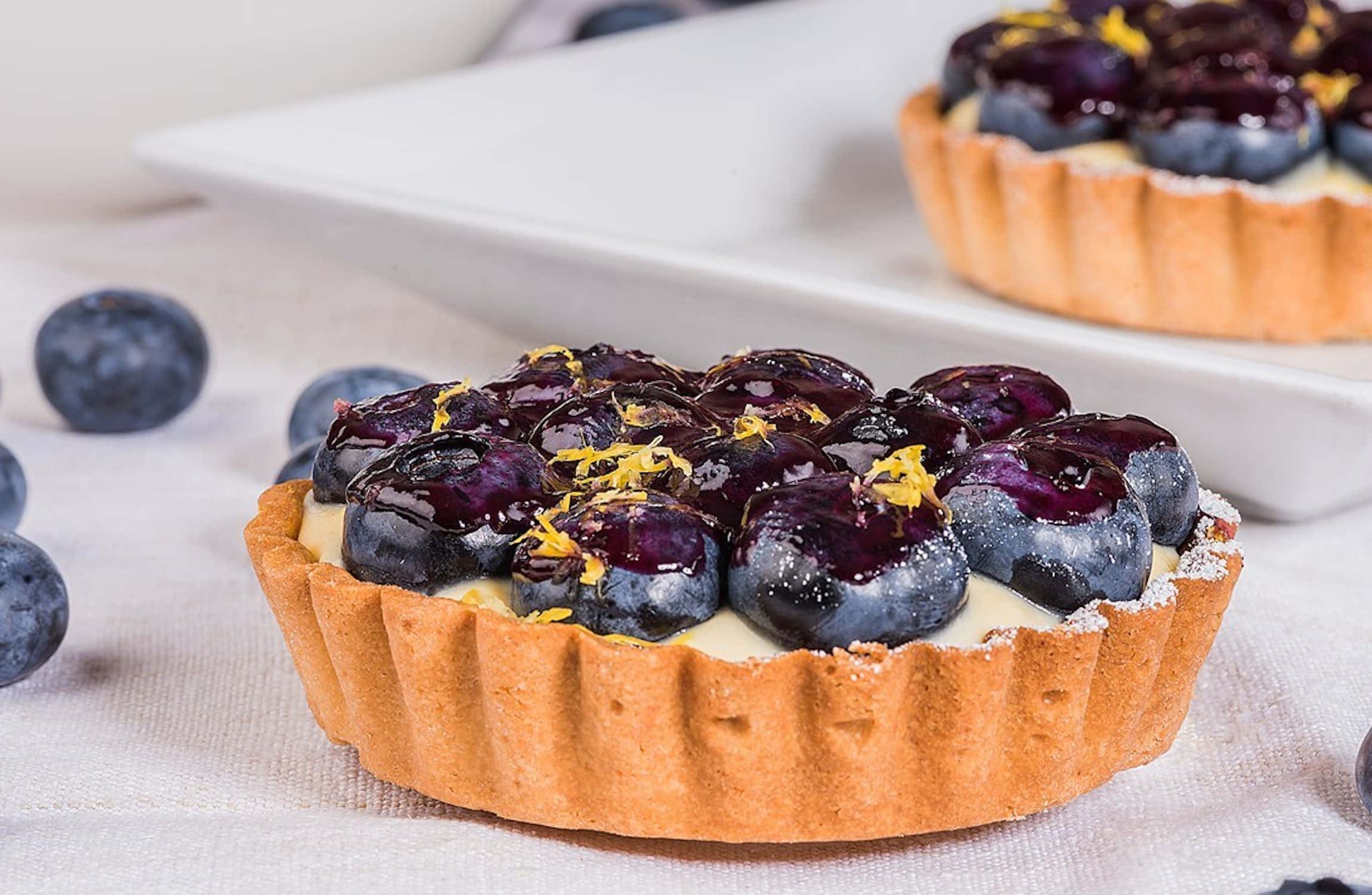 Blueberries Tart 桌布 壁紙 — 金黃色塔的近距離特寫，內餡充滿奶油卡士達，上面鋪滿閃亮藍莓和檸檬皮。