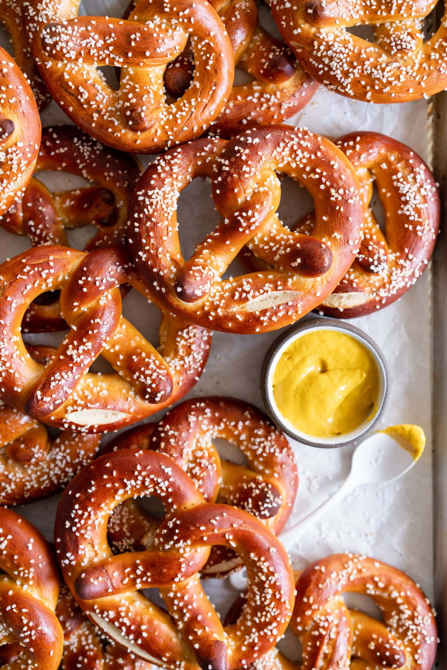 Pretzel 桌布 壁紙 — 金黃色椒鹽卷餅撒上粗鹽，旁邊放著一小碗奶油黃色芥末醬，置於烘焙紙上。