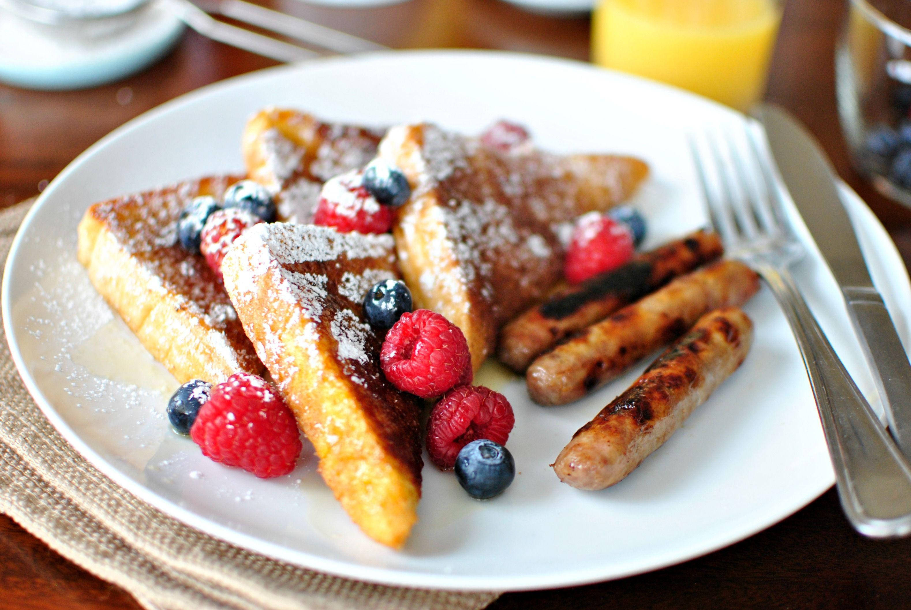 French Toast 桌布 壁紙 — 金黃色法式吐司三角形，撒上糖粉，搭配新鮮覆盆子和藍莓，放在白色盤子上，附有烤香腸。