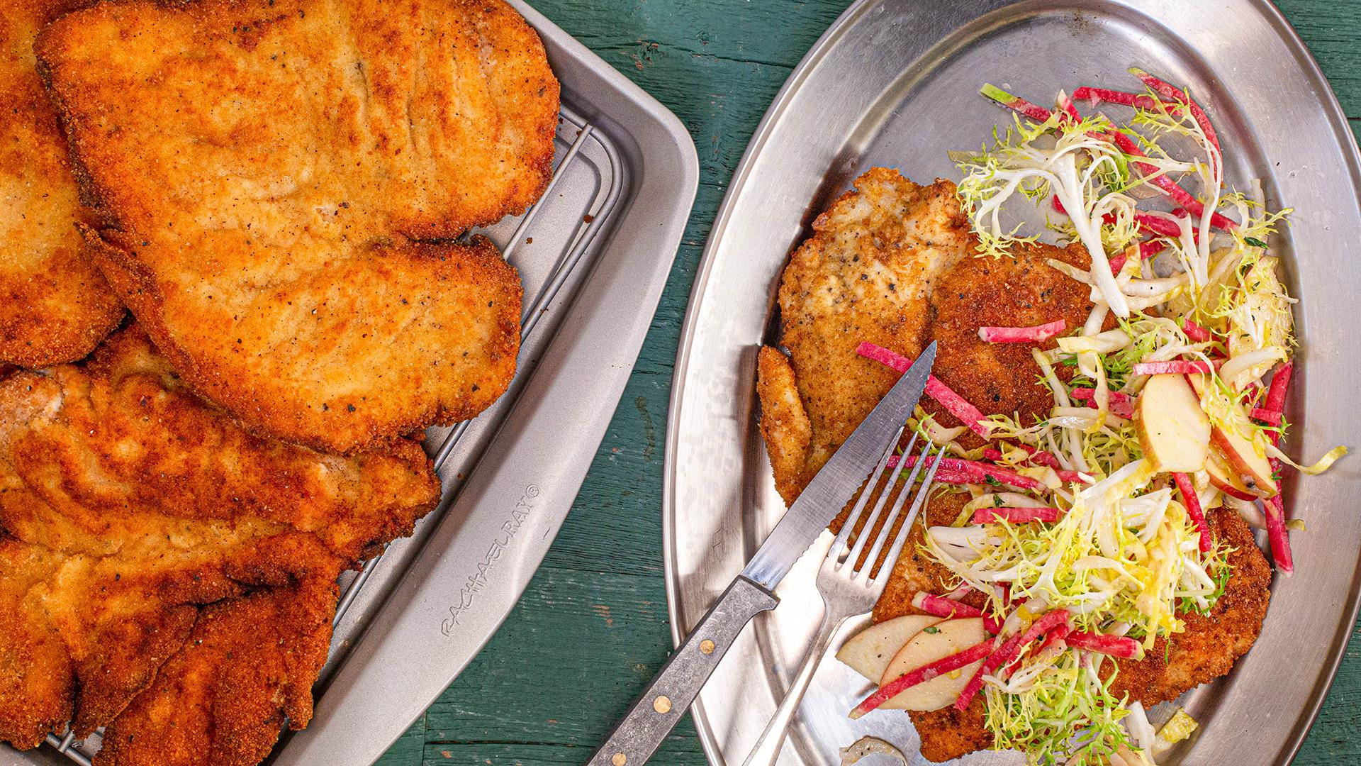 Lamb 桌布 壁紙 — 金黃色炸肉排（Schnitzel）熱騰騰地放在托盤上，搭配色彩繽紛的苦苣沙拉，置於鄉村風格的綠色木桌上。
