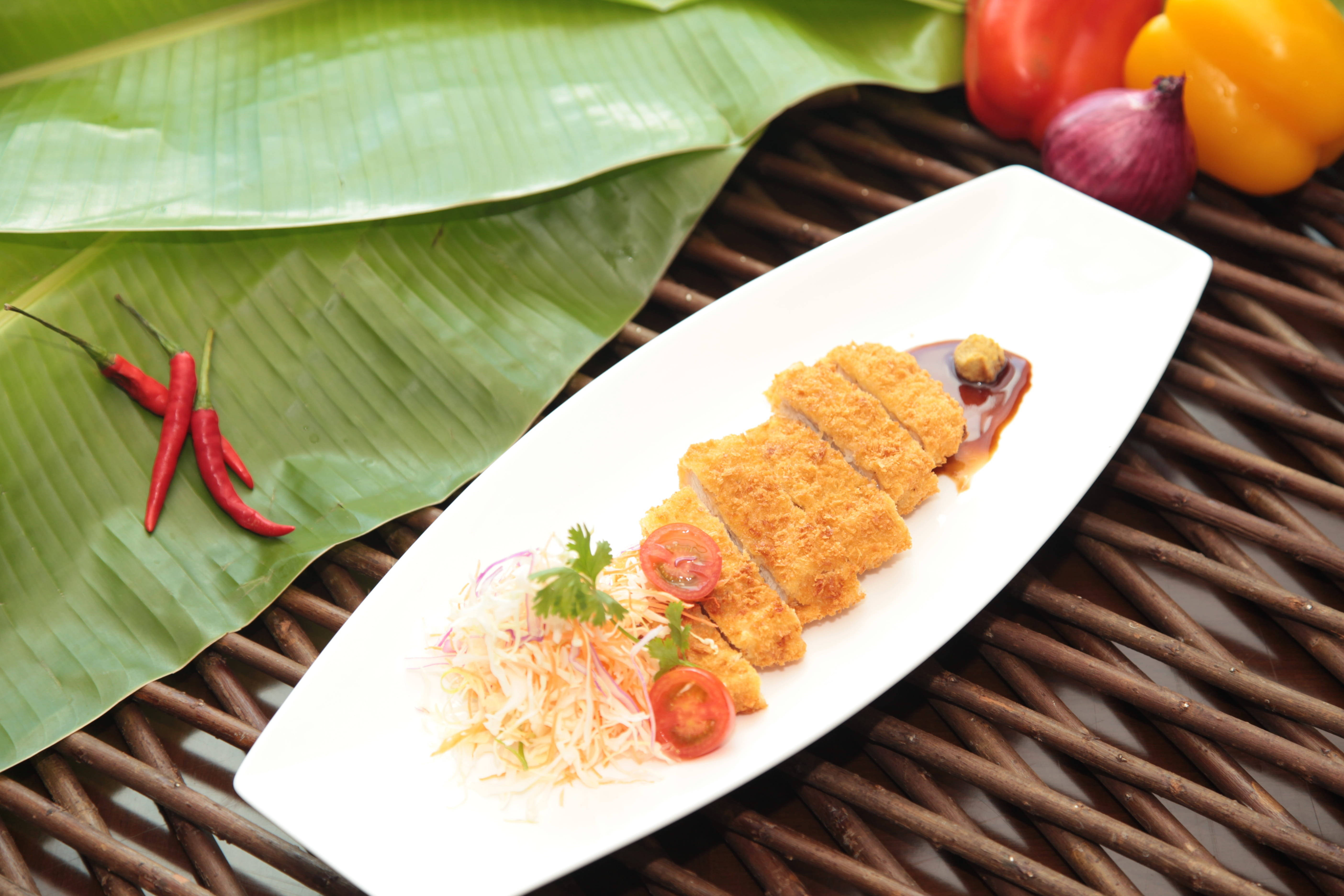 Food Photography 桌布 壁紙 — 金黃色炸豬排切片放在白色盤子上，配上醃漬蔬菜和醬汁，旁邊有香蕉葉和新鮮辣椒。