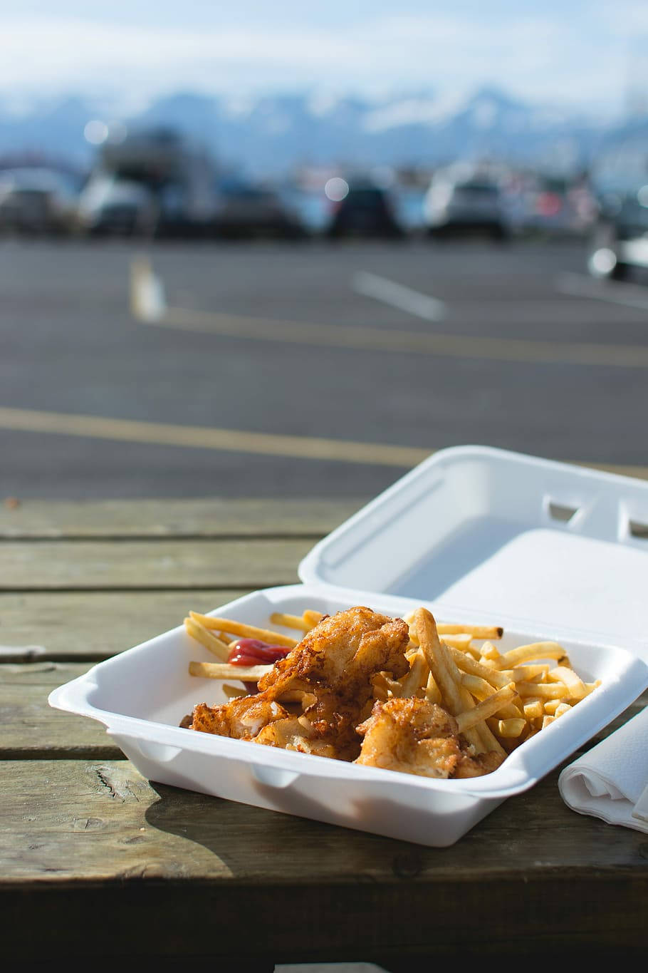 fast food 桌布 壁紙 — 金黃色炸雞和薯條放在白色外帶盒中，置於一張風化的木製野餐桌之上，背景是模糊的雪山。