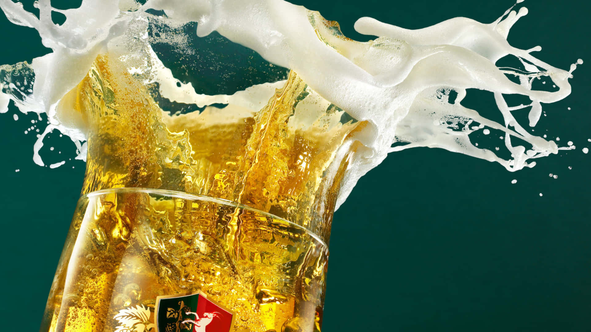 Beer 桌布 壁紙 — 金黃色的啤酒從玻璃杯中噴湧而出，帶有泡沫豐富的白色泡沫，在深藍綠色背景上戲劇性地飛濺。