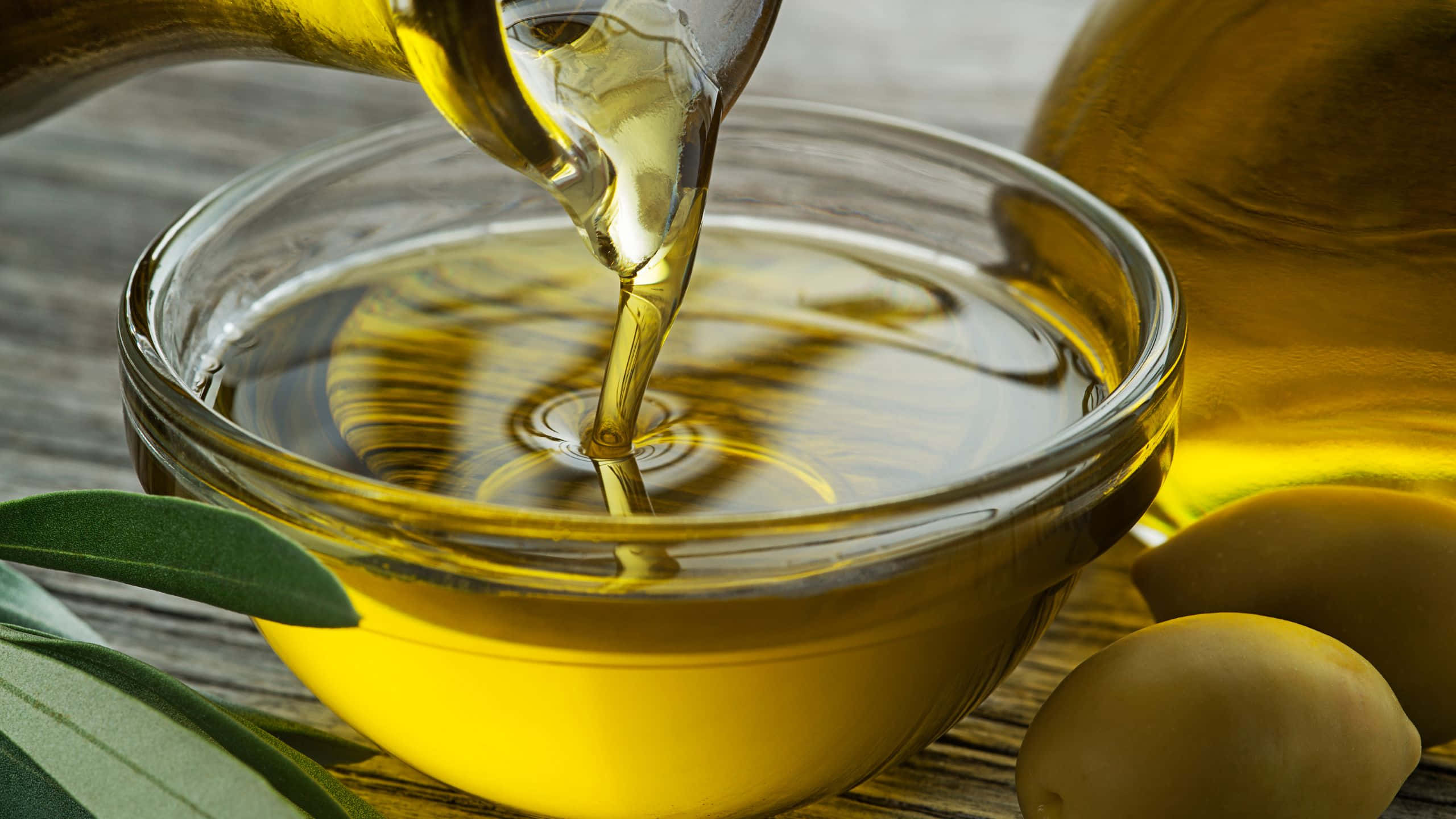Oil 桌布 壁紙 — 金黃色的橄欖油以慢動作倒入玻璃碗中，旁邊是新鮮的橄欖和綠葉，放在質樸的木質表面上。