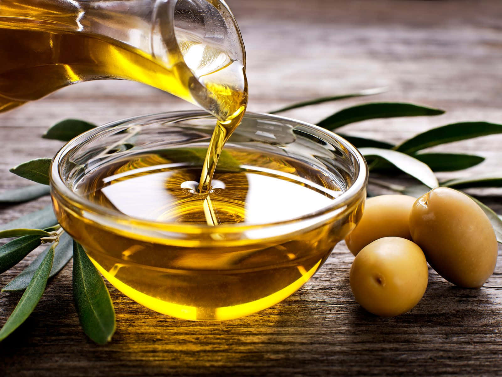 Oil 桌布 壁紙 — 金黃色的橄欖油順暢地倒入一個玻璃碗中，旁邊是新鮮的橄欖和橄欖葉，置於質樸的木質表面上。