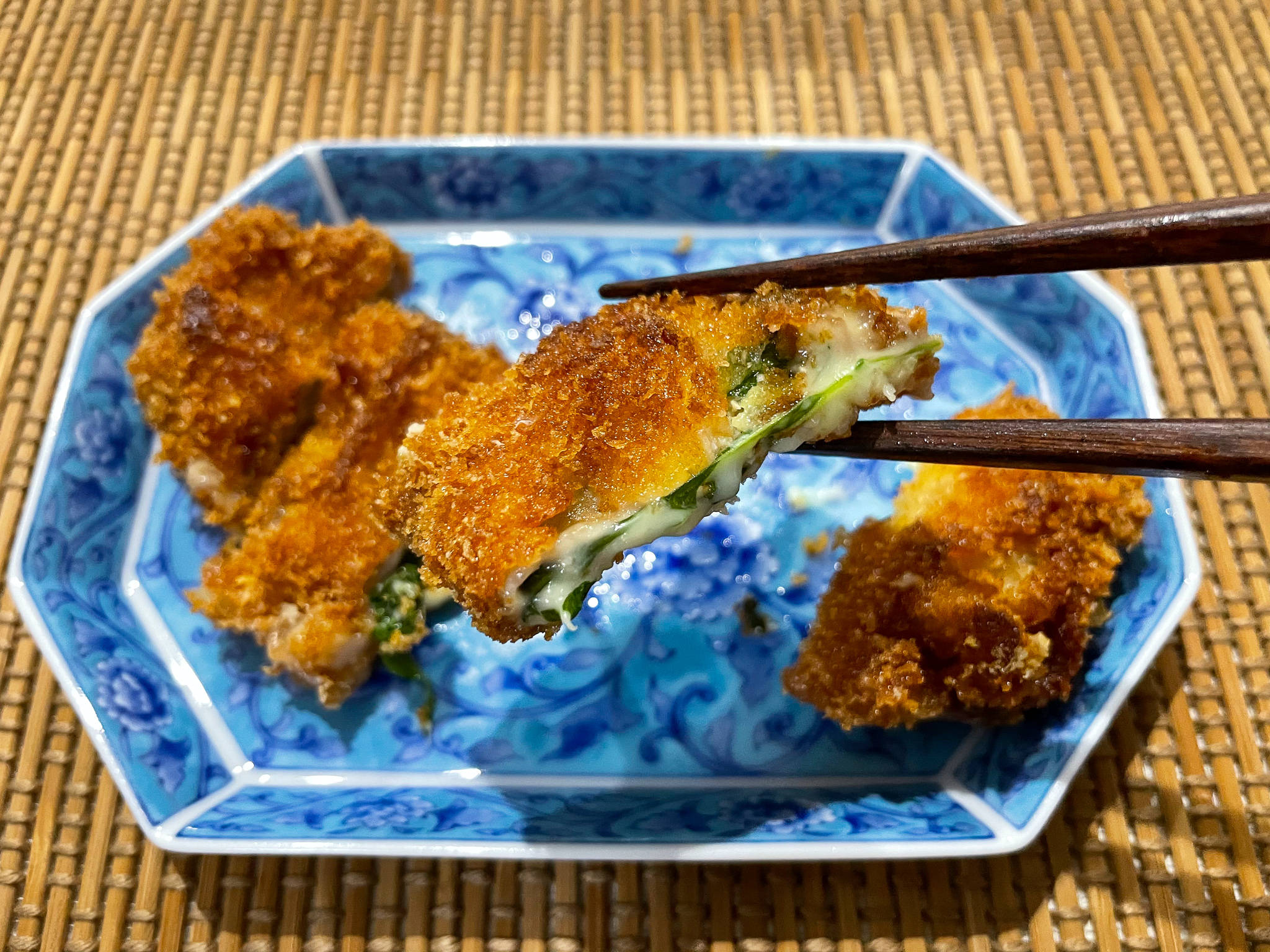 Food Photography 桌布 壁紙 — 金黃色的炸雞塊，內有綠色餡料，用筷子夾起，放在竹蓆上的藍色圖案盤子上。