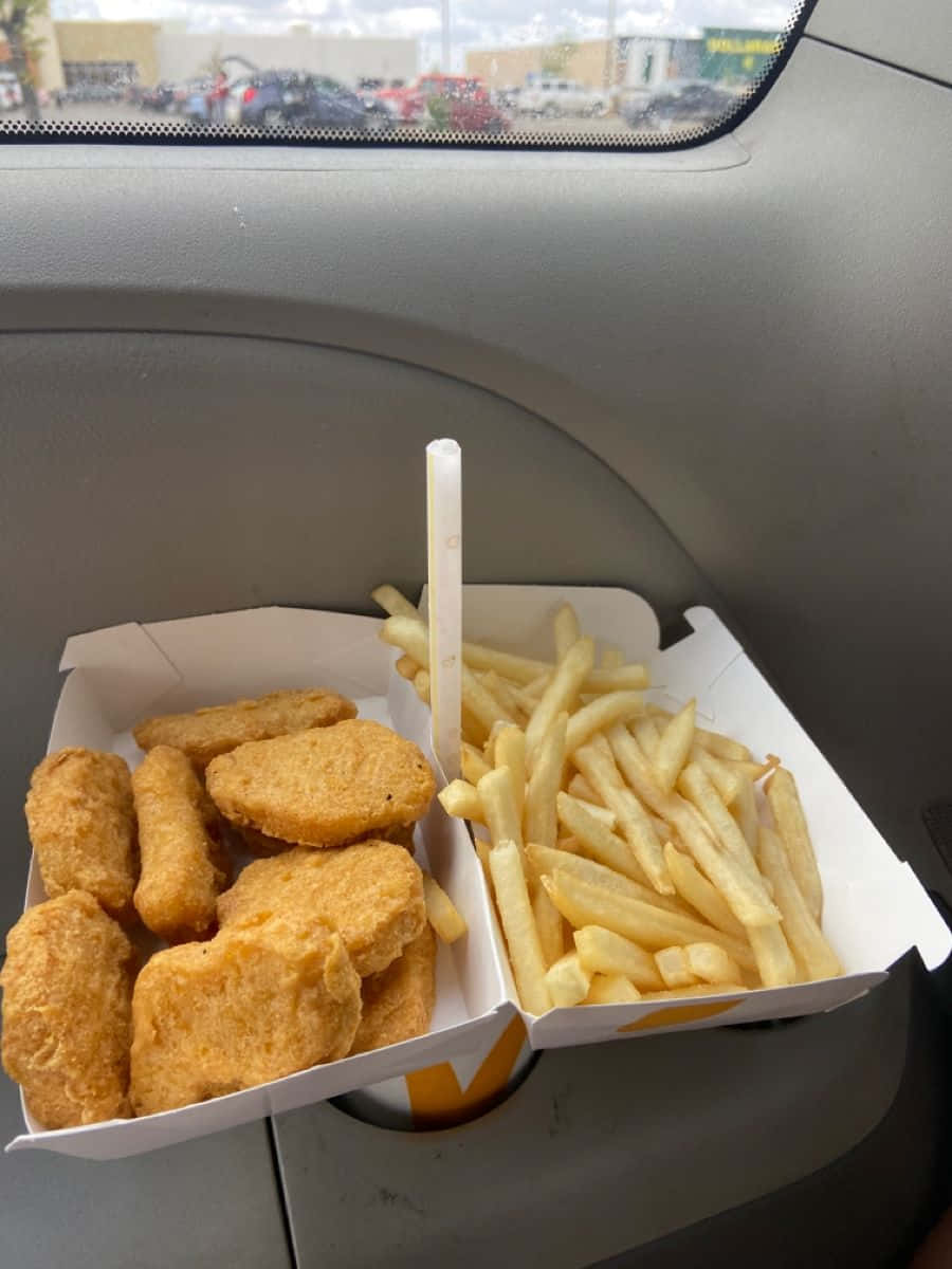 麥當勞 桌布 壁紙 — 金黃色的炸雞塊和薯條放在白色盒子裡，擱在汽車杯架上，可看到停車場景色。