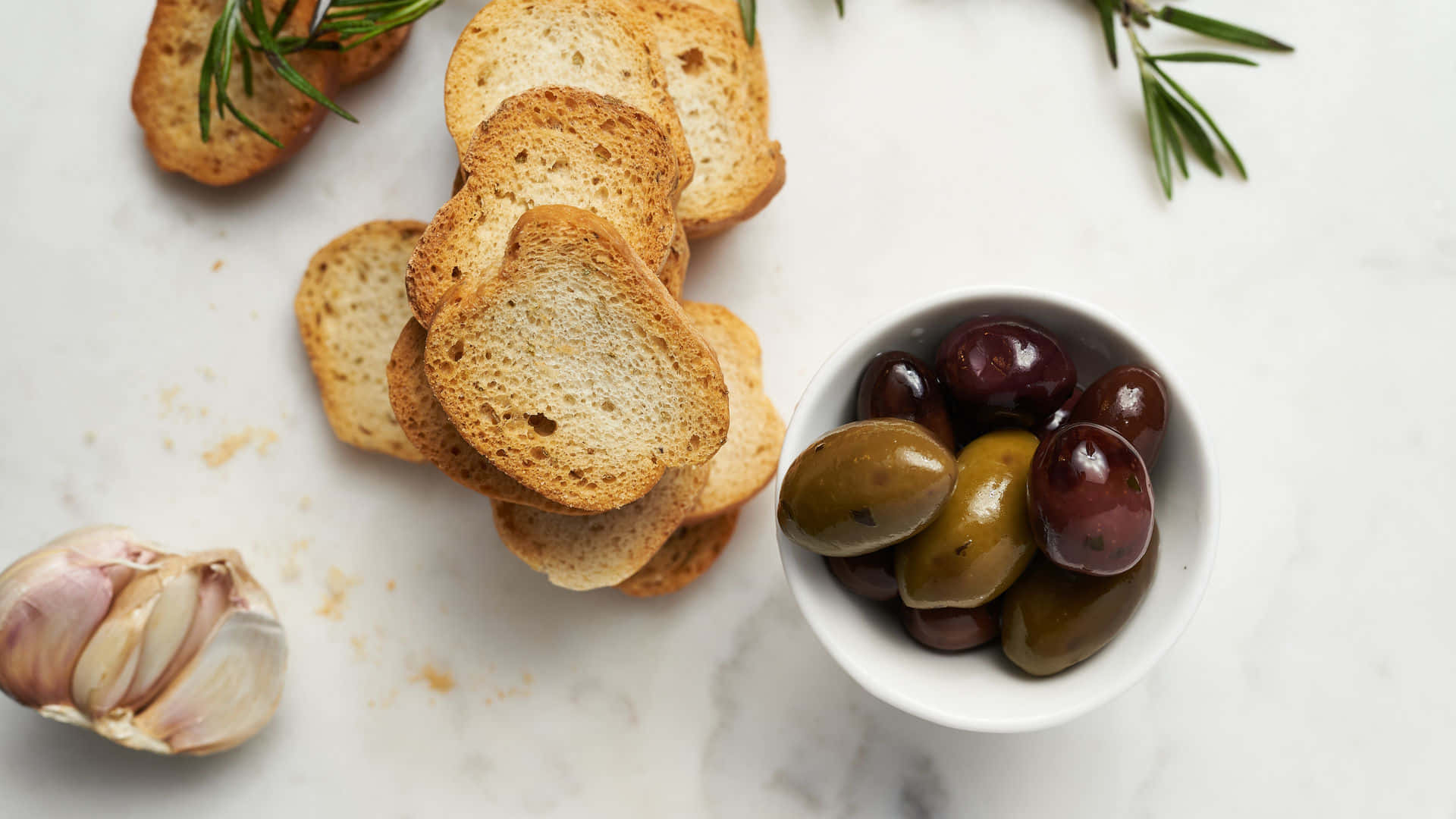 Bread 桌布 壁紙 — 金黃色的烤麵包片和一碗光澤的黑橄欖和青橄欖放在白色大理石表面上，配以新鮮迷迭香和大蒜。