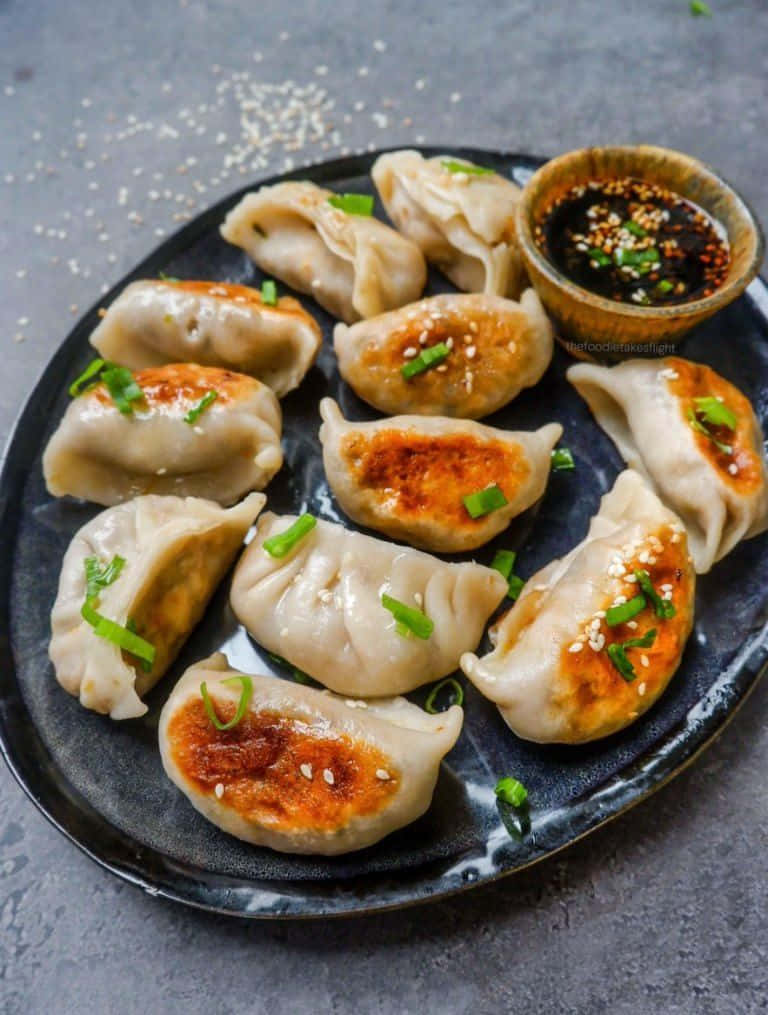 Local Food 桌布 壁紙 — 金黃色的煎餃，上面撒上蔥花和芝麻，放在深色盤子上，旁邊配上蘸醬。