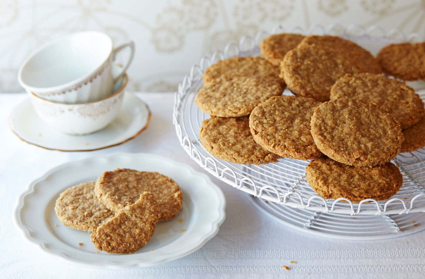 Biscuit 桌布 壁紙 — 金黃色的燕麥餅乾放在白色金屬架上和小碟子旁邊，旁邊是堆疊的復古茶杯，桌子鋪著蕾絲桌布。