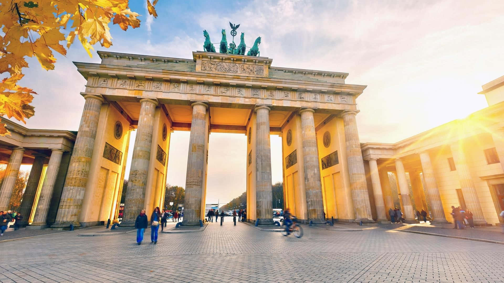 Berlin 桌布 壁紙 — 金黃色的秋葉框住了標誌性的布蘭登堡門，沐浴在溫暖的日落光線中，人們在其宏偉的柱廊間漫步。