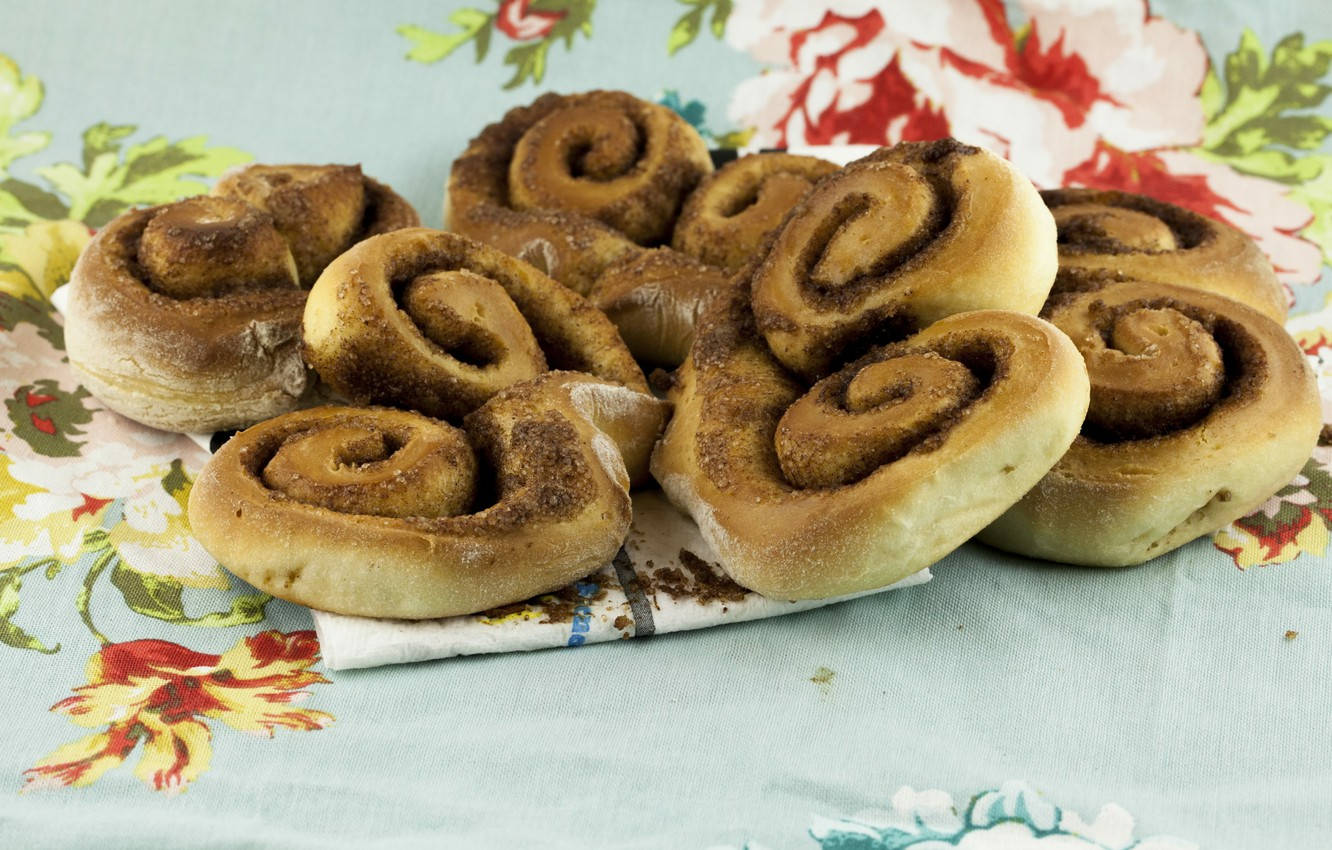 Pastry 桌布 壁紙 — 金黃色的肉桂捲帶有深色漩渦，放在花卉圖案的布上。